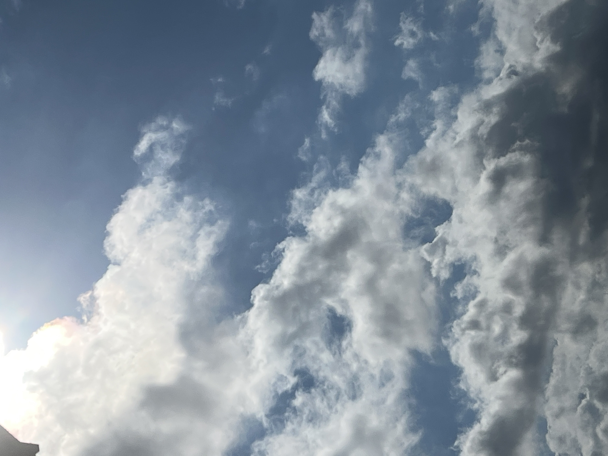 The image is a photograph of a cloudy sky, taken from an outdoor perspective. The sky is a mix of bright blue and white, with various cloud formations. The sun is partially obscured by a large, white cloud in the upper left quadrant of the image, with a rainbow-like shimmer visible around its edges. The clouds are dense and appear to be moving, creating a dynamic and textured sky. The overall lighting suggests it is daytime, likely with some sunlight breaking through the clouds. There is no indication of any specific city or location within the image.
