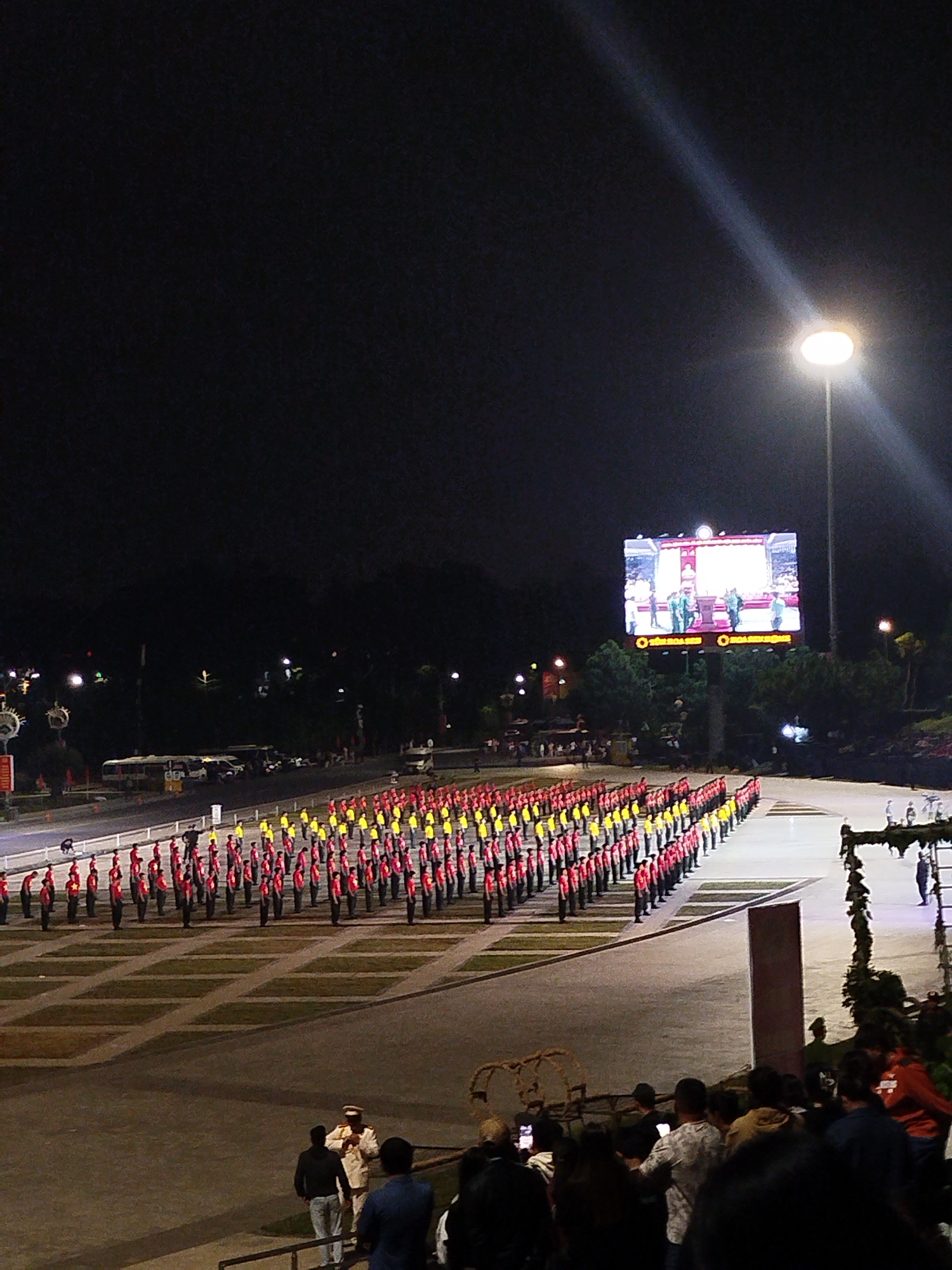 The image depicts a nighttime outdoor ceremony or performance at the Lam Vien Square in Da Lat, Vietnam. A large crowd of spectators is gathered in the foreground, observing a formation of individuals in uniform on a paved area. The uniformed individuals, many wearing red and yellow attire, are arranged in neat rows, suggesting a synchronized performance or parade. In the background, a large LED screen displays an image of people in uniform, and beyond that, trees and buildings are visible, illuminated by artificial lights. A prominent streetlight casts a bright beam across the dark sky. The overall atmosphere appears to be one of a formal event or celebration.