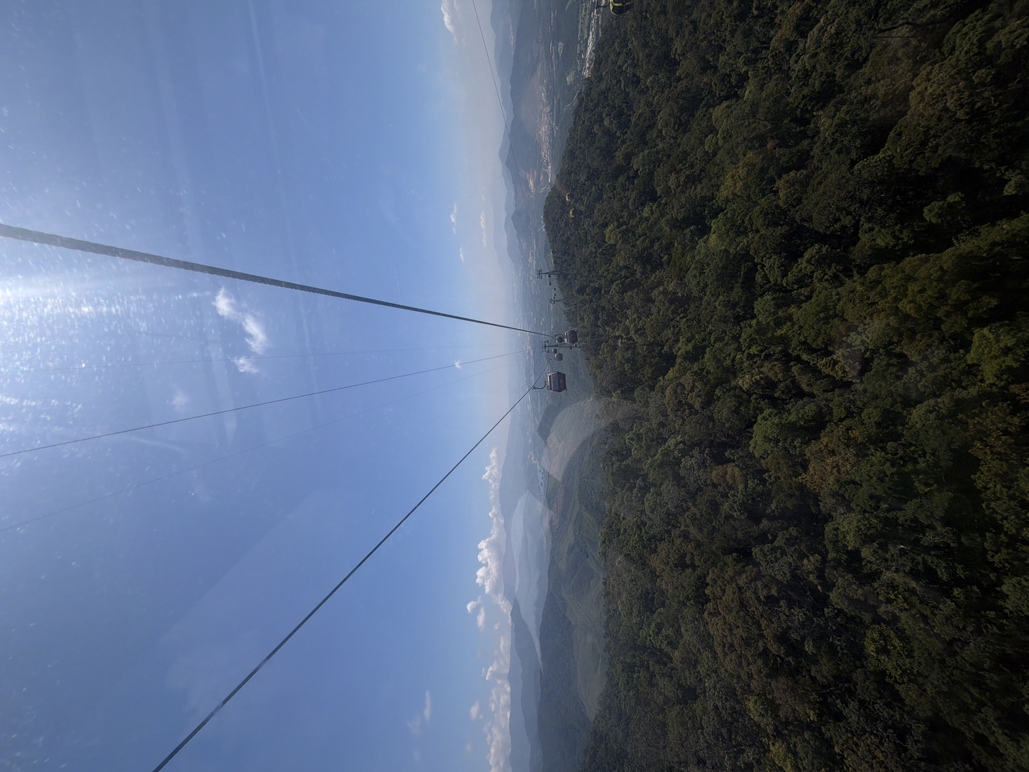 The image captures a view from a cable car in Đà Nẵng, Vietnam. The scene shows the cable lines stretching across the frame, with cable cars suspended. To the right is a dense forest covering a hillside. In the distance, hazy mountains are visible. The sky is blue with scattered clouds. There are no people visible within the frame.