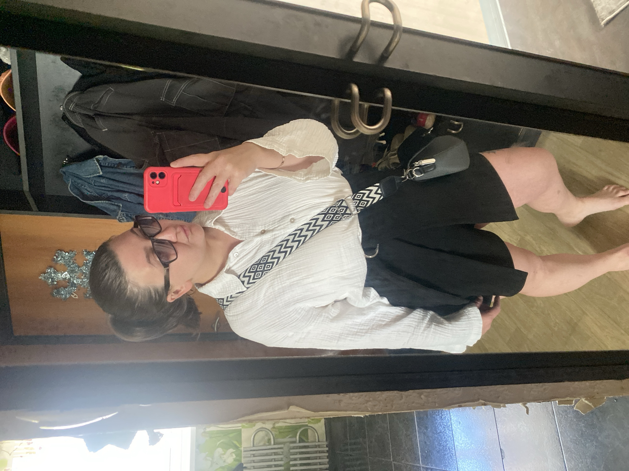 A woman stands in front of a mirror in what appears to be a closet. She is wearing a white textured shirt, black shorts, and sunglasses. She has a red phone in her right hand and a black purse with a patterned strap over her left shoulder. In the background, there are clothes hanging on a rack, and hats on a shelf. The floor is wooden. The mirror reflects a section of the room, showing a window with light-colored curtains and a tiled floor with a patterned rug. The scene suggests a personal space, possibly a bedroom or dressing area, located in a private residence in Chaikovsky, Russia.