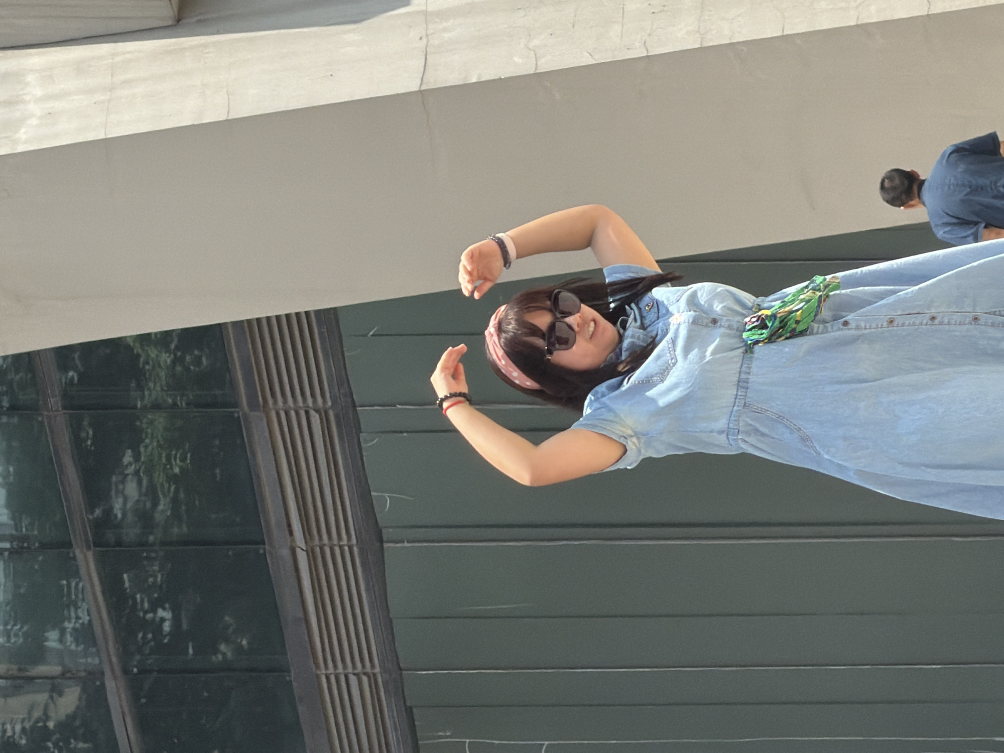 A young Asian woman, wearing a light blue denim dress, a pink polka dot headband, and sunglasses, stands with her arms raised and hands near her head, facing forward. Her dress is buttoned up the front and has a small, colorful fabric accessory tied around the waist. To the right and lower in the frame, a man in a dark blue shirt is visible from behind, also facing away from the camera. The background consists of a dark green wall with vertical lines and a light-colored concrete structure. Above the green wall are windows with horizontal blinds, reflecting trees. The scene appears to be outdoors in a modern urban environment, possibly in Yuzhong District, China.