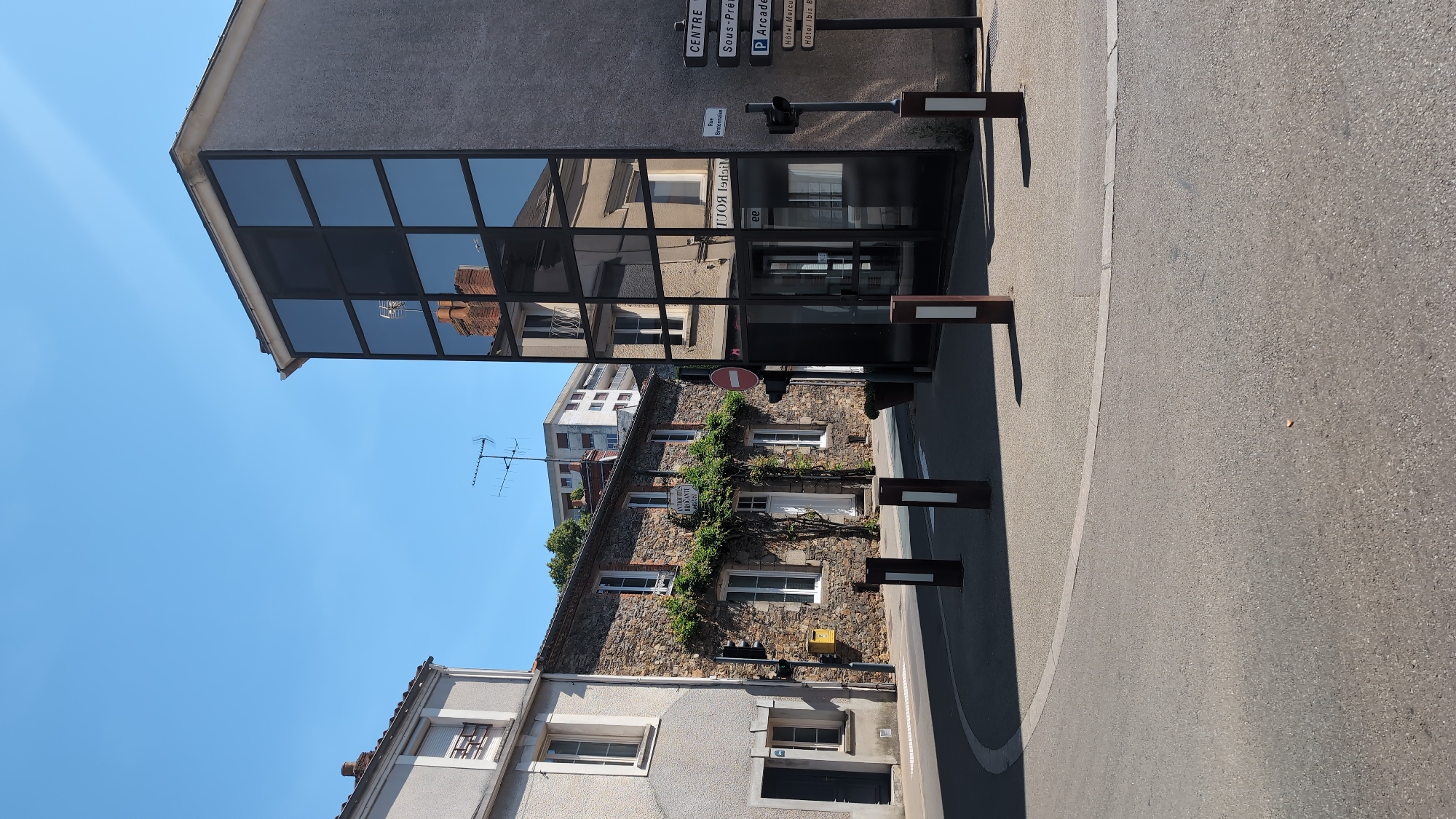 The image depicts a street scene in Cholet, France, likely at an intersection. On the right is a modern building with a facade of large, dark-tinted glass panels, reflecting the surrounding environment, including a stone building with white-framed windows and a chimney. A street signpost on this building indicates directions to "CENTRE", "Sous-Préf.", "P Arcade", "Hôtel Mercu.", and "Hôtel Ibis B.". Adjacent to this modern building, to the left, is a traditional stone building with green foliage climbing its facade, white-trimmed windows, and a white door. Above this stone building, a taller, multi-story building with white balconies is visible, along with antennas on its roof. A grey, two-story building with a white-trimmed window on the upper floor and a dark door on the lower floor stands to the far left. The street is paved with asphalt and a circular grey area with white lines. Several bollards are positioned in the foreground. The sky is clear and blue. No people are visible in the frame.