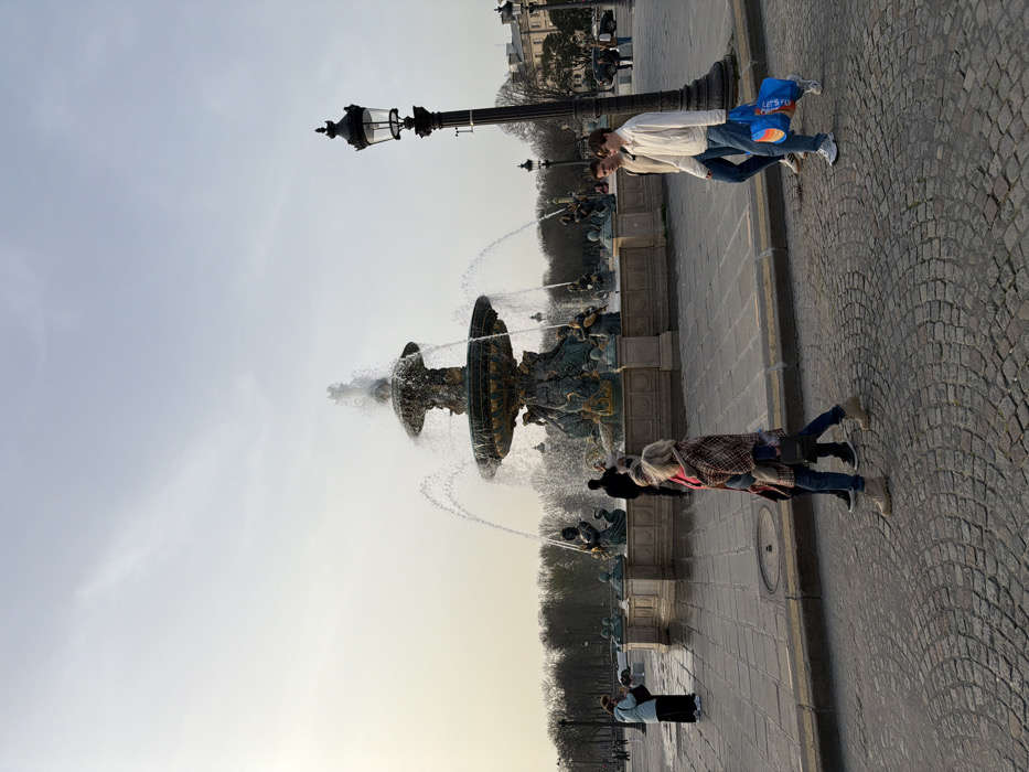 The image depicts an outdoor urban scene in the Place de la Concorde, Paris, France. The central feature is a large, ornate, multi-tiered bronze and stone fountain, identified as one of the twin fountains in the square (likely the Fontaine des Mers), actively spraying water from its upper and lower basins. The fountain is surrounded by sculpted figures, including mermaids and tritons.
In the foreground, a blonde-haired individual, wearing a plaid jacket/shawl, red top, blue jeans, and light-colored boots, walks from left to right across a cobblestone street, carrying a dark bag. To their right, two young men also walk left to right. The lead man wears a light-colored jacket, blue jeans, and white sneakers, carrying a blue "Let's Fly" branded tote bag. The second man wears a light brown jacket, blue jeans, and white sneakers.
Further back on the left, a person in a light blue jacket and dark pants stands near the fountain's edge, holding a smartphone, seemingly capturing an image. Another person in dark attire is partially visible near the fountain's base, walking away from the camera.
A tall, black, ornate streetlamp with a single glass lantern fixture stands prominently on the right side of the frame, separating the cobblestone area from the paved plaza around the fountain. The background features bare trees, indicating a season outside of full foliage, and distant classical Parisian buildings under a pale, clear sky. The ground consists of cobblestone paving in the foreground, transitioning to larger square pavers around the fountain.