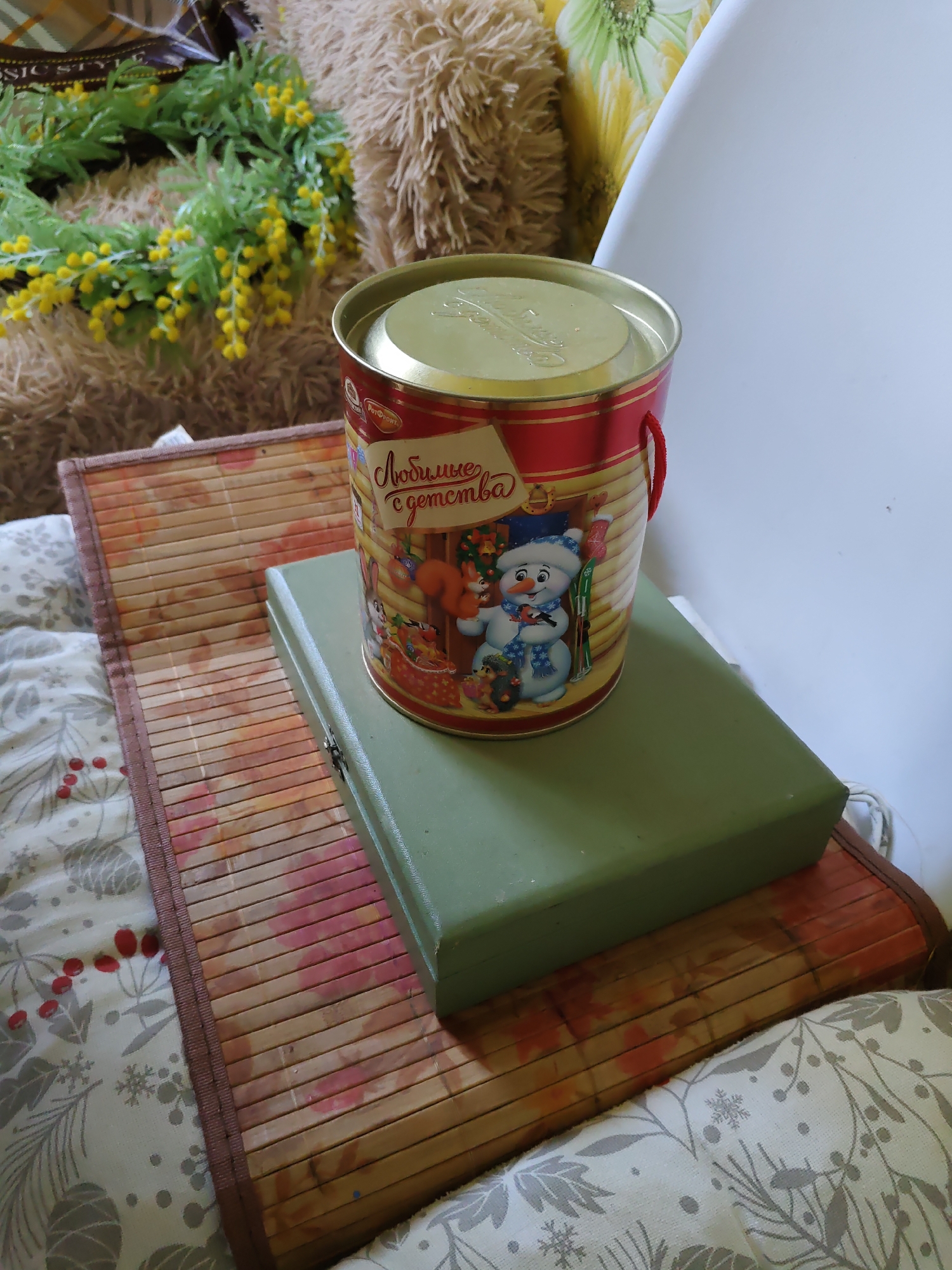 A tin of sweets with a picture of a snowman is placed on a green box, which is on a bamboo mat. The bamboo mat is on a patterned fabric surface. In the background, there is a fluffy object and a wreath of green leaves and yellow flowers. The scene appears to be indoors, possibly in a home, and the overall impression is one of seasonal decoration or a gift display. No specific location in Rostov-on-Don is identifiable.