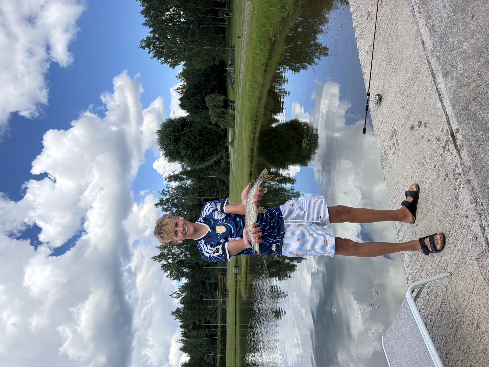 A young man stands on a concrete platform next to a body of water, holding a freshly caught fish. He is wearing a blue and white soccer jersey and striped shorts with small yellow duck figures. He has blonde hair and is wearing white earbuds. To his right, a fishing rod with a reel rests on the concrete. The water reflects the cloudy blue sky and the surrounding trees. In the background, there is a grassy area with trees and some structures, suggesting a park or recreational area. The location is identified as Mežpļavas, Latvia.