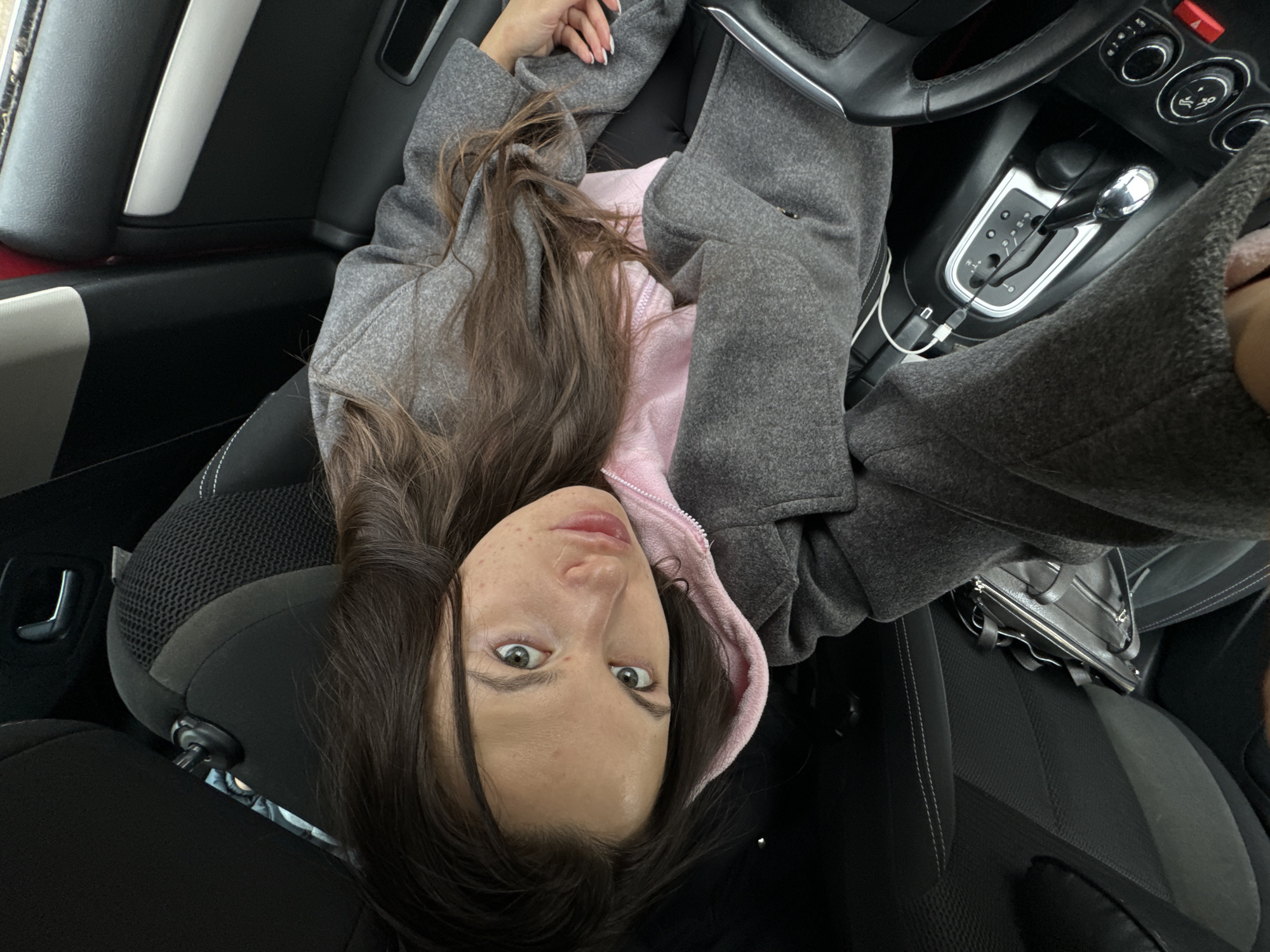 A woman with long brown hair, wearing a pink hooded sweatshirt and a gray coat, is sitting in the driver's seat of a car. The car's interior is visible, including the steering wheel, dashboard controls, gear shift, and seats. A black purse is on the passenger seat. The location is Unknown, Unknown.