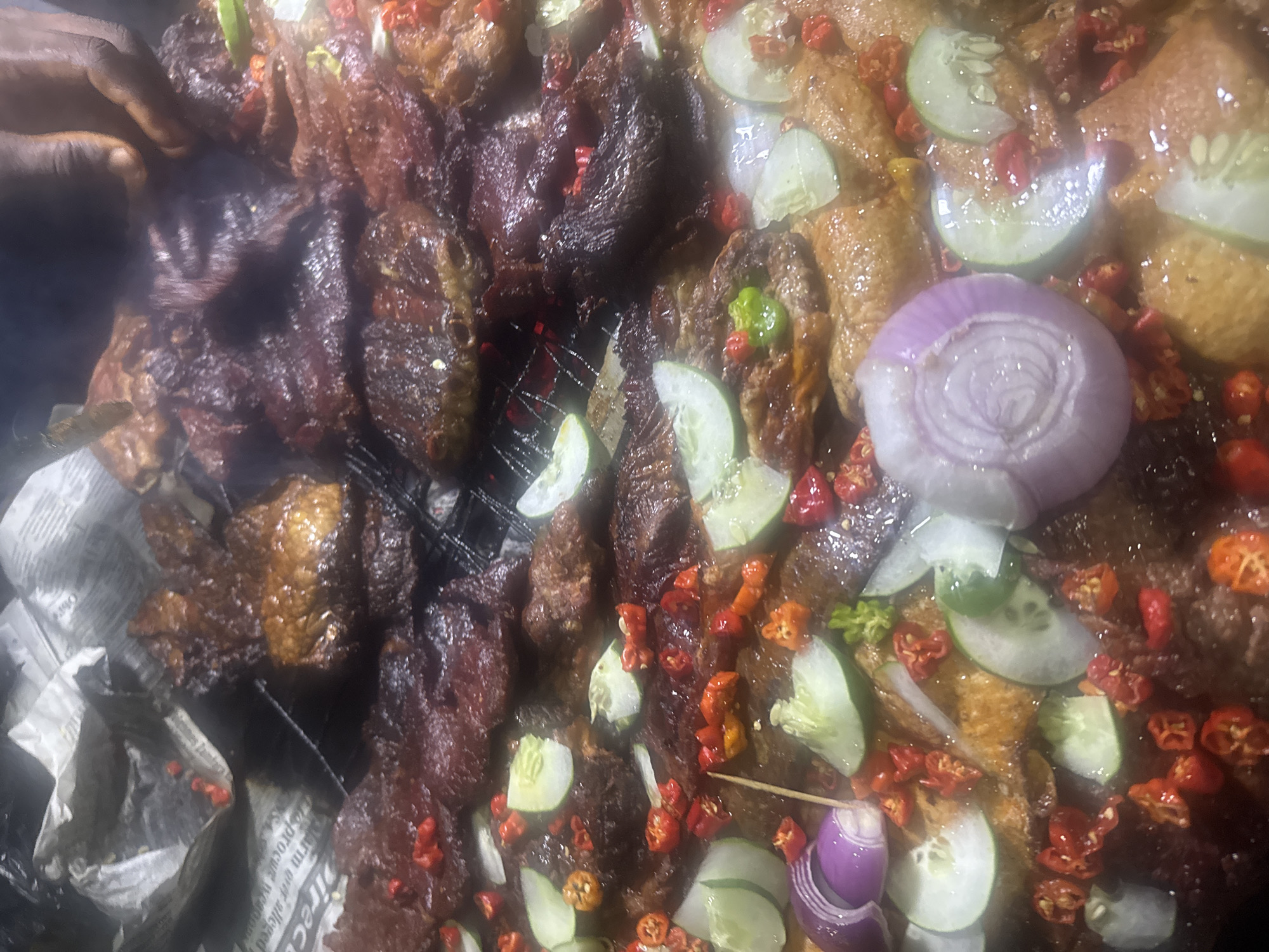 A close-up, overhead shot displays a large quantity of grilled meat, possibly beef, arranged on a black metal grill. The meat is dark brown and appears to be seasoned with spices. Scattered across the meat are slices of cucumber, chopped red chilies, and slices of red onion. Some of the meat is slightly charred. The background is out of focus and appears to be a surface covered with what looks like newspapers. The image evokes a street food vendor setting, potentially in a bustling market area in Abuja, Nigeria, given the context provided. The lighting is warm and appears to be artificial, possibly from overhead lights or cooking fires. The overall impression is one of a vibrant and flavorful dish being prepared or served.