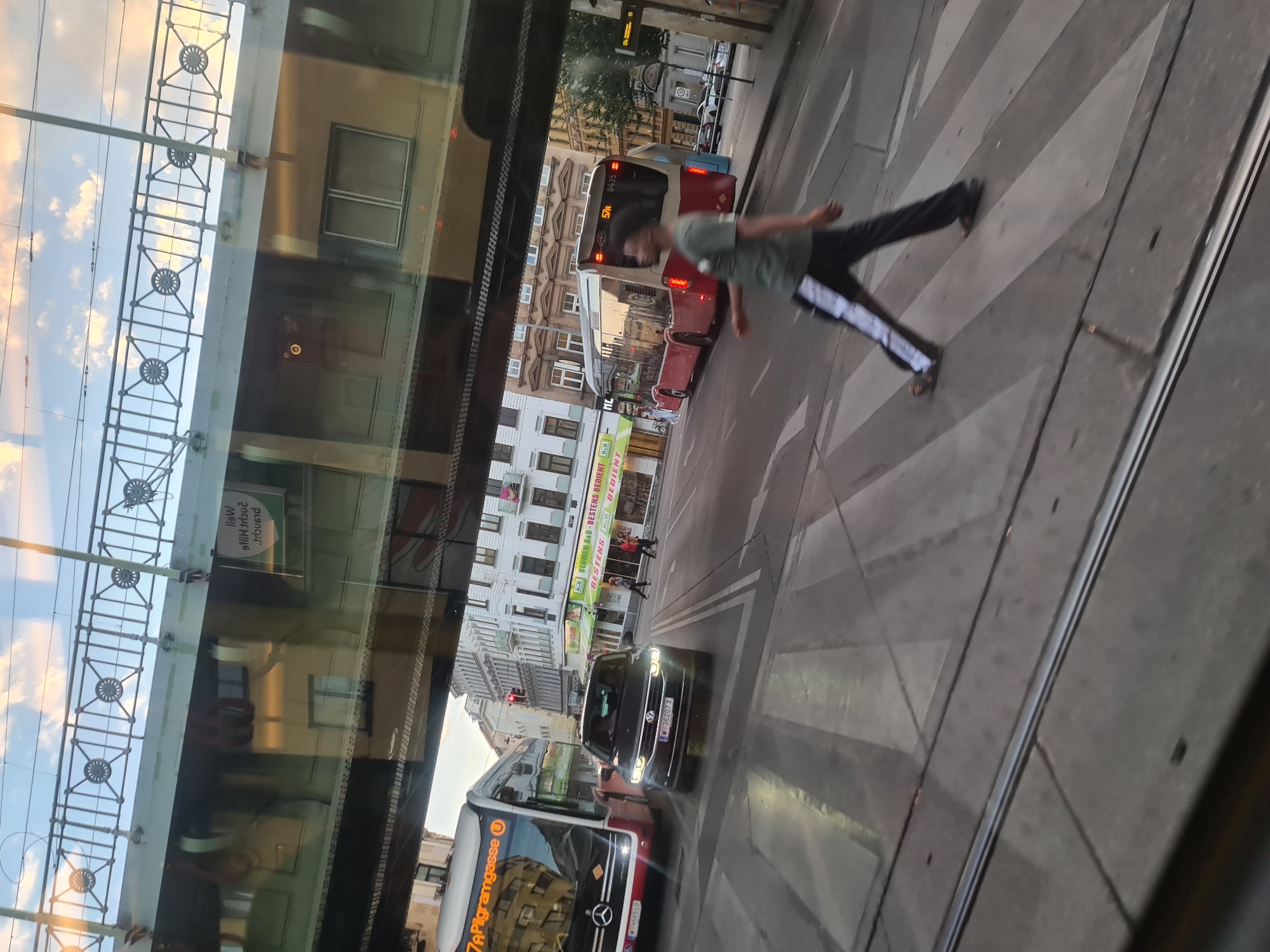 The image captures a street scene in Vienna, Austria, viewed from a moving vehicle, likely a tram or bus, as indicated by the reflection of the sky and an overhead structure. The foreground is dominated by tram tracks embedded in the asphalt, with a pedestrian crossing marked by white stripes.
A young Black man is walking across the pedestrian crossing from left to right, wearing a green t-shirt and dark trousers with white stripes. He is blurred due to motion.
In the middle ground, a dark-colored Volkswagen Tiguan SUV is stopped at the crossing, facing the camera. Its headlights are on, and the license plate is partially visible. To the left of the SUV, a red and white public transport bus, identified by the sign "7A Pilgramgasse U", is also stopped. Further down the street, another similar red and white bus is visible.
The background consists of multi-story buildings lining the street, exhibiting typical European architecture. Shops with awnings are visible at street level. The sky is a mix of blue with scattered white clouds, with the warm tones of sunset visible, suggesting the image was taken in the late afternoon or early evening. The overall impression is a busy urban environment during daylight hours.