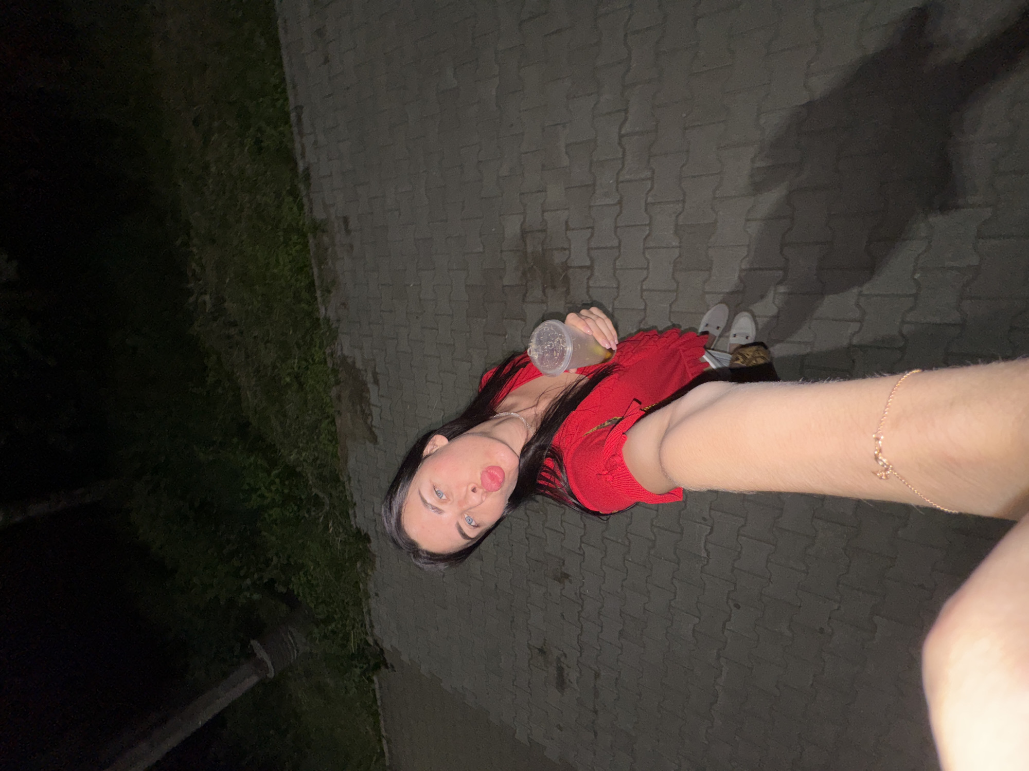 A woman with dark hair and blue eyes is taking a selfie at night. She is wearing a red top and a necklace. She is holding a drink in a plastic cup and has a bracelet on her arm. She is standing on a paved walkway, with grass and trees in the background. A street light is visible on the left side of the image. The scene appears to be outdoors, possibly in a park or on a street in the city of Chaikovsky, Russia.