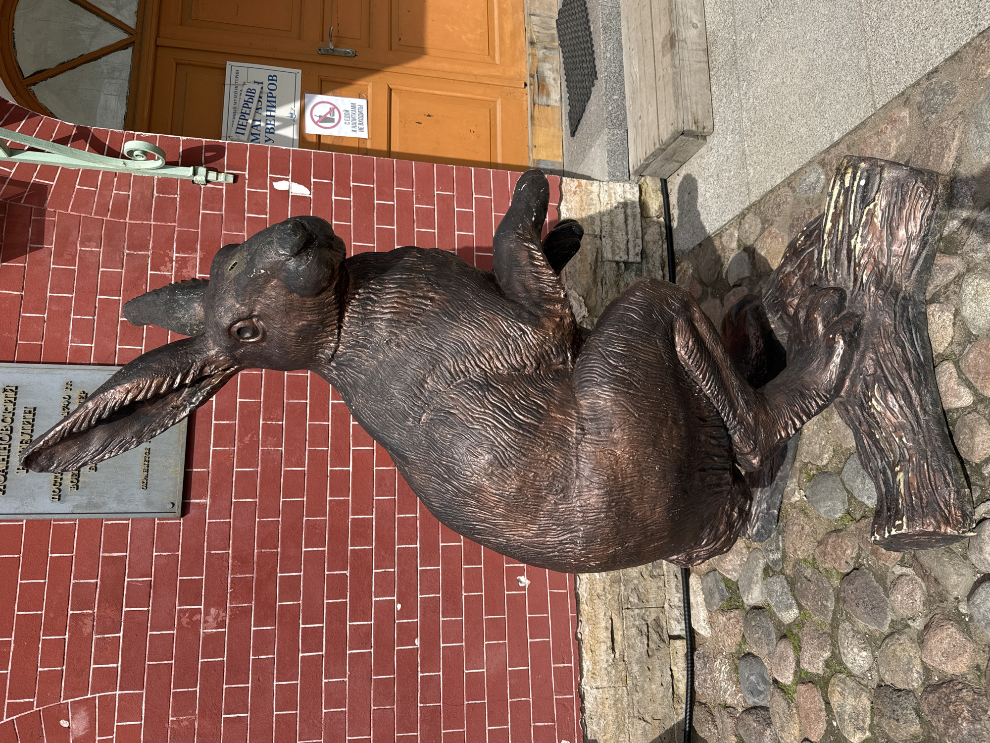 A large, dark bronze statue of a rabbit is depicted sitting on cobblestone pavement. The rabbit is positioned upright, facing left, with its front paws raised and its head slightly tilted. Its textured surface mimics fur, and it appears to be resting against a bronze tree stump.
The background to the left of the rabbit features a red brick wall with white mortar. Attached to this wall is a metal plaque with partially visible Russian text, including "ДОАННОВСКИЙ", "ТАМЕЛИН", "1740", and "МУЗЕЙ ИСТОРИИ" (Museum of History), suggesting a historical context. To the right of the rabbit, an old, light brown wooden door is visible, featuring an arched window detail at the top. On the door, white signs in Russian read "ПЕРЕРЫВ" (Break) and "МАГАЗИН УВЕНИРОВ" (Souvenir Shop). A prominent sign below these, accompanied by a universal "no food or drink" icon, states "С ЕДОЙ И НАПИТКАМИ НЕ ВХОДИТЬ!" (Do not enter with food and drinks!).
The ground beneath the statue and extending into the foreground is covered with varied grey and brown cobblestones. The scene is brightly lit by natural sunlight, indicating daytime and clear weather, with shadows cast across the pavement. This outdoor setting is located in Saint Petersburg, Russia.