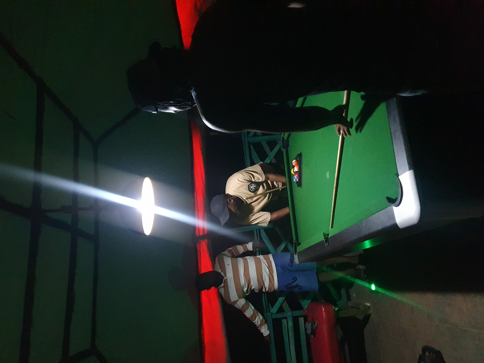 Three individuals are gathered around a pool table in a dimly lit, outdoor setting, likely a bar or recreational area, in Lokoja, Nigeria. The table, covered in green felt, has a rack of colored balls and a cue stick positioned for play. One person, wearing a striped polo shirt and blue shorts, stands to the left, leaning on a railing. Another individual, in a tan shirt and baseball cap, is bent over the pool table, holding a cue. A third person, silhouetted in the foreground, faces away from the camera, also positioned near the pool table. The environment appears to be covered, with a sloped, dark green ceiling supported by dark beams, and red fabric accents visible in the background. A bright overhead light casts strong beams and shadows, illuminating parts of the scene. A green laser light beam is visible on the floor near the pool table.