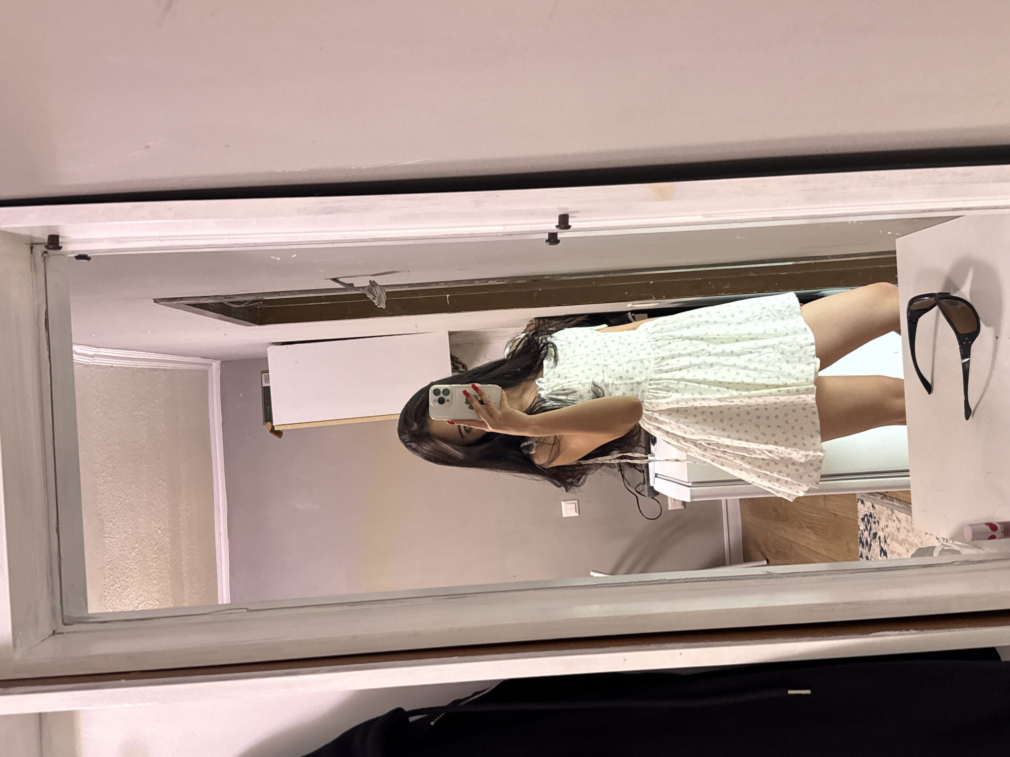 A young woman with long, dark hair is taking a mirror selfie in a room. She is wearing a light-colored, floral-print dress and is holding a smartphone in her right hand. A pair of sunglasses rests on a white surface in the foreground. The room appears to be a bedroom or dressing area, with a closet to the left, a mirror on the right, and furniture and walls visible in the reflection. The overall impression is of a casual, indoor setting in a residential space, possibly an apartment or house. Based on the provided information, the location is Sary-Oy, Kyrgyzstan.
