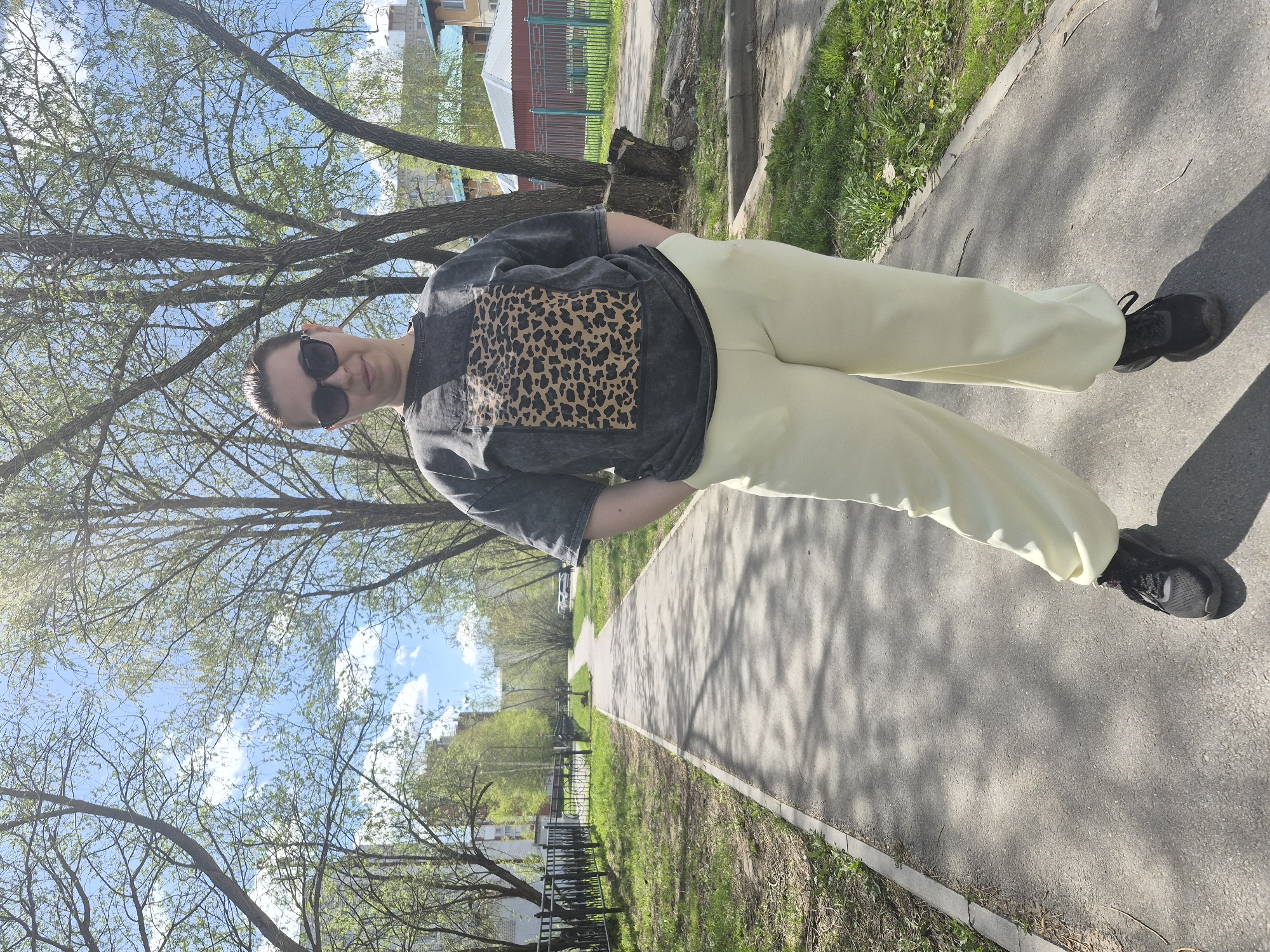 A full-body shot depicts a woman standing on a sidewalk, presumably in Tobolsk, Russia. She wears a gray, short-sleeved t-shirt with a leopard print square on the front, paired with light yellow pants and dark sneakers. Sunglasses obscure her eyes. The background reveals a paved path, green grass, trees with emerging foliage, and glimpses of residential buildings with colorful fencing. The sky is visible through the tree branches, showing a mix of blue sky and white clouds.