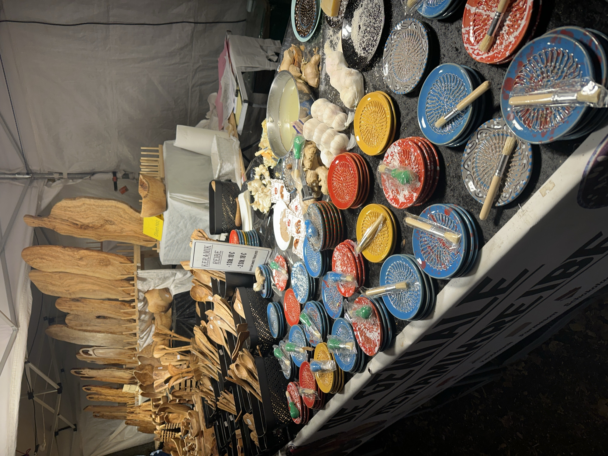 The image shows a stall at a Christmas market in Bruchhausen-Vilsen, Germany, likely at the "Bruchhausen-Vilsener Weihnachtsmarkt". The stall displays a variety of wooden kitchen utensils and ceramic graters. In the background, numerous wooden items, possibly decorative or functional pieces, are arranged on shelves. In the foreground, stacks of colorful ceramic graters, primarily in red, yellow, blue, and grey, are laid out on a table covered with a black cloth. Each grater is accompanied by a small brush. There are also some garlic bulbs and ginger root on display. A sign on the stall reads "KERAMIK REIBE" and pricing information in German. The market appears to be outdoors, under a tent, and it is nighttime, indicated by the artificial lighting. No people are clearly visible in the frame.