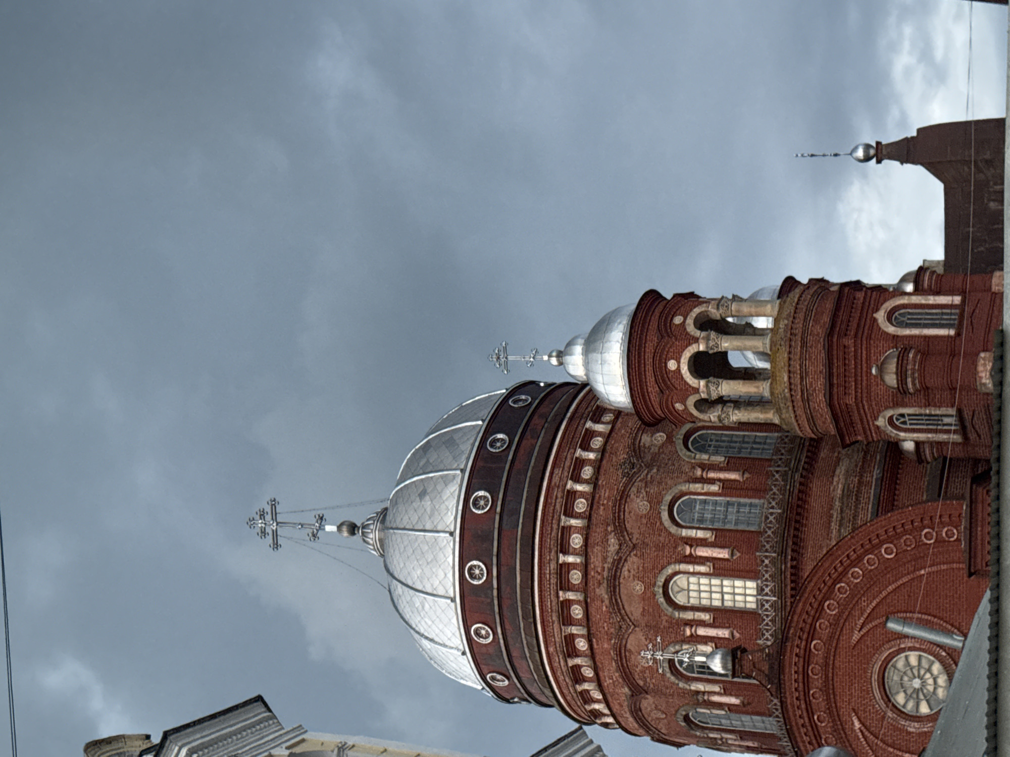 The image shows the Ascension Cathedral in Urzhum, Russia, under a cloudy sky. The cathedral is constructed of red brick and features multiple domes, including a large central dome with a silver, segmented surface topped by a cross. Smaller domes and decorative elements are visible on other sections of the building. The architecture includes arched windows, decorative stonework, and tiered sections. The scene is a low-angle shot, emphasizing the height and grandeur of the cathedral against the overcast sky. There are no people visible in the image.