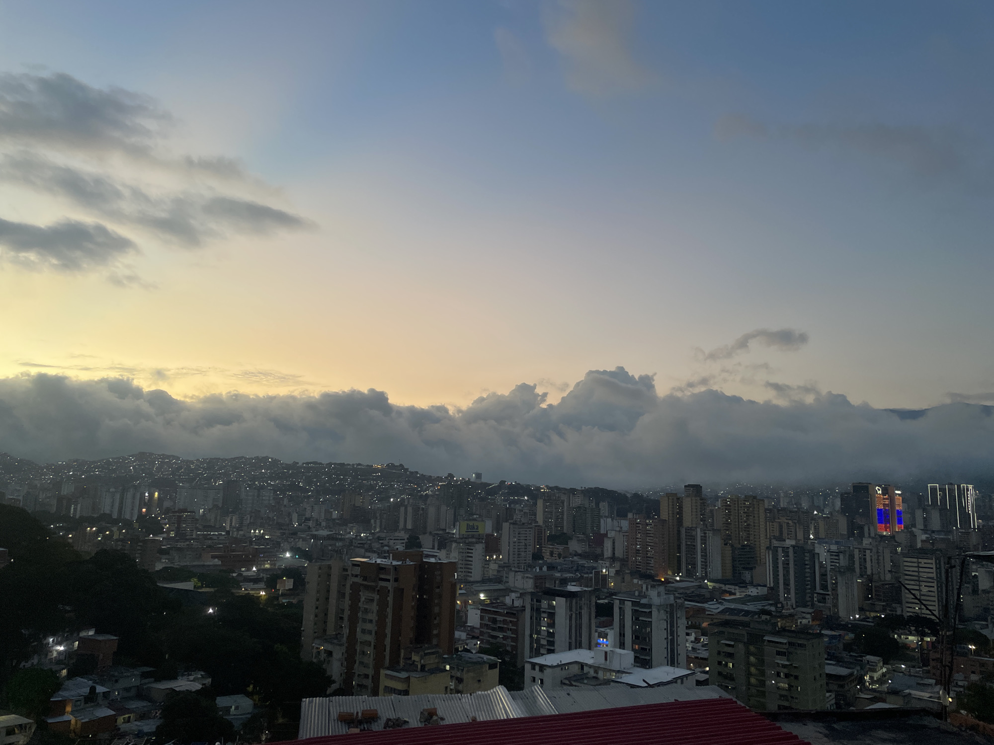 The image shows a high-angle view of the city of Caracas, Venezuela, at dusk. The foreground contains parts of buildings with a red rooftop visible. In the mid-ground, a dense urban landscape is made up of various high-rise buildings. A layer of clouds and haze obscures parts of the city and stretches across the horizon, blending into the sky, which transitions from light blue to yellow near the horizon, indicating either sunrise or sunset. The buildings in the background include some with lit windows. The towers with blue, yellow, and red colors represent the colors of the Venezuelan flag.