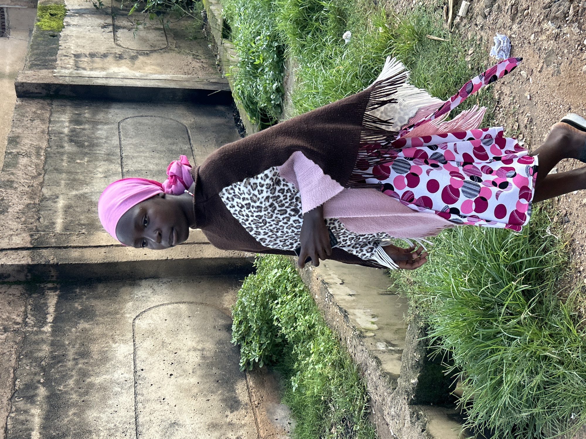 In Jos, Nigeria, a young Black girl with a pink headscarf and a pink and white circular patterned dress walks past a concrete wall and overgrown grass. She is wearing a brown poncho over a white top with a leopard print design and pink trim. Her dress has a fringe hem. She is holding a dark object in her right hand, and her left hand is partially visible. The background consists of a textured concrete wall, a ditch filled with lush green grass, and a dirt path with some debris.