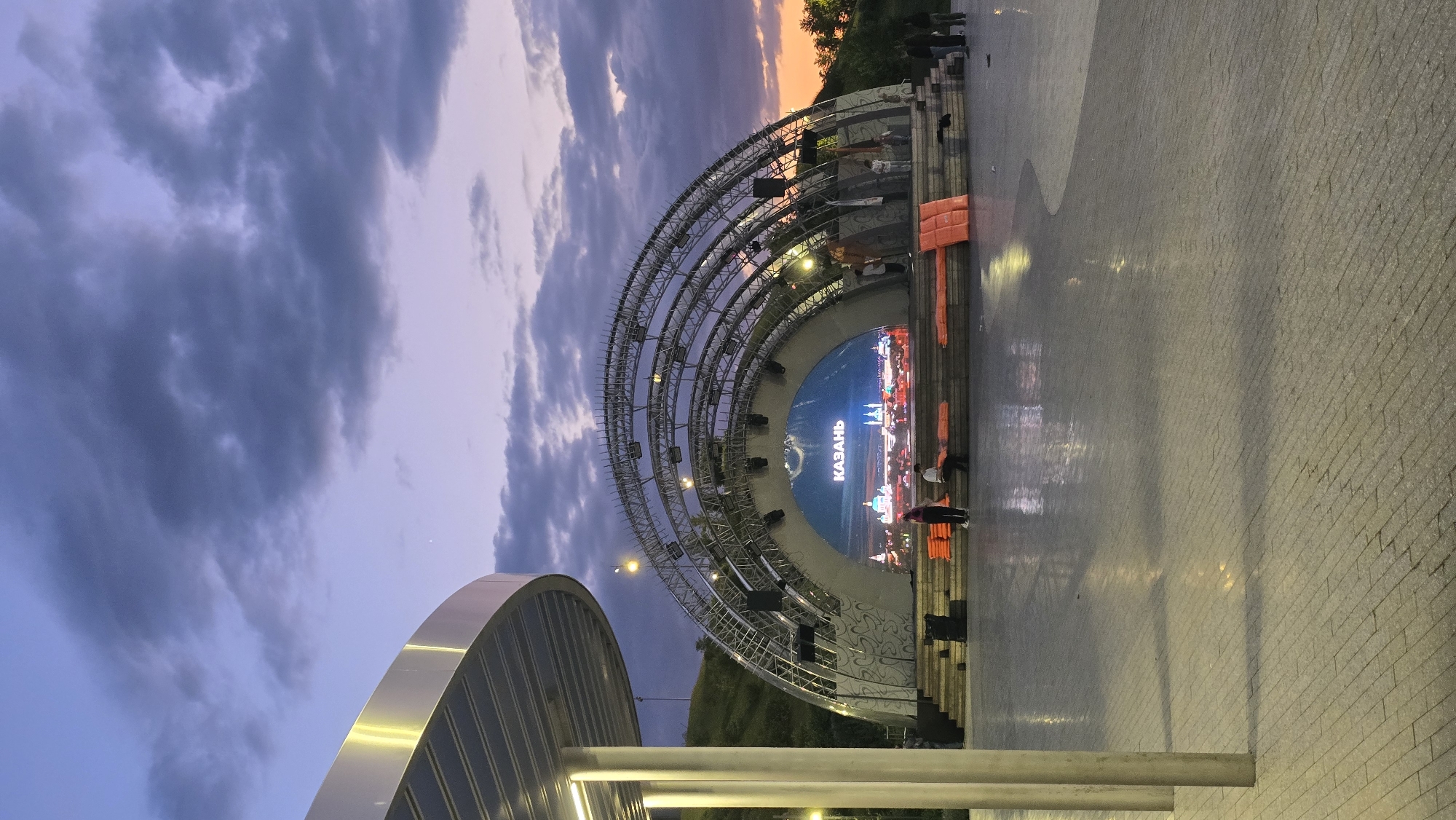 This image captures an outdoor stage area in Kazan, Russia, at dusk. The central feature is a large, semi-circular architectural steel truss structure forming a stage backdrop. This structure incorporates a prominent curved LED screen displaying a vibrant, illuminated cityscape, with the word "КАЗАНЬ" (KAZAN) in white Cyrillic script clearly visible. The stage itself consists of multiple tiers of steps, upon which several rectangular orange objects, resembling cushions or safety gear, are arranged.
In the left foreground, a modern metallic canopy with a curved, reflective roof and two cylindrical support columns is partially visible. The paved ground surface in the foreground and mid-ground is visibly wet, reflecting the stage lights and overhead ambient illumination.
Approximately seven individuals are observed in the scene. Two are seated on the lower steps of the stage, one in light-colored attire and another in dark. A third person, wearing a pink top and dark trousers, stands near them. On the right side of the stage, a group of four individuals is positioned, some standing, with one wearing a light-colored head covering.
The sky exhibits twilight colors, transitioning from soft lavender and deep blue hues overhead to a subtle orange glow on the far right horizon. Scattered, dark grey clouds are present across the sky, and a faint star is visible in the upper left quadrant. A grassy hill rises behind the stage on the left, while trees are silhouetted against the horizon on the right.