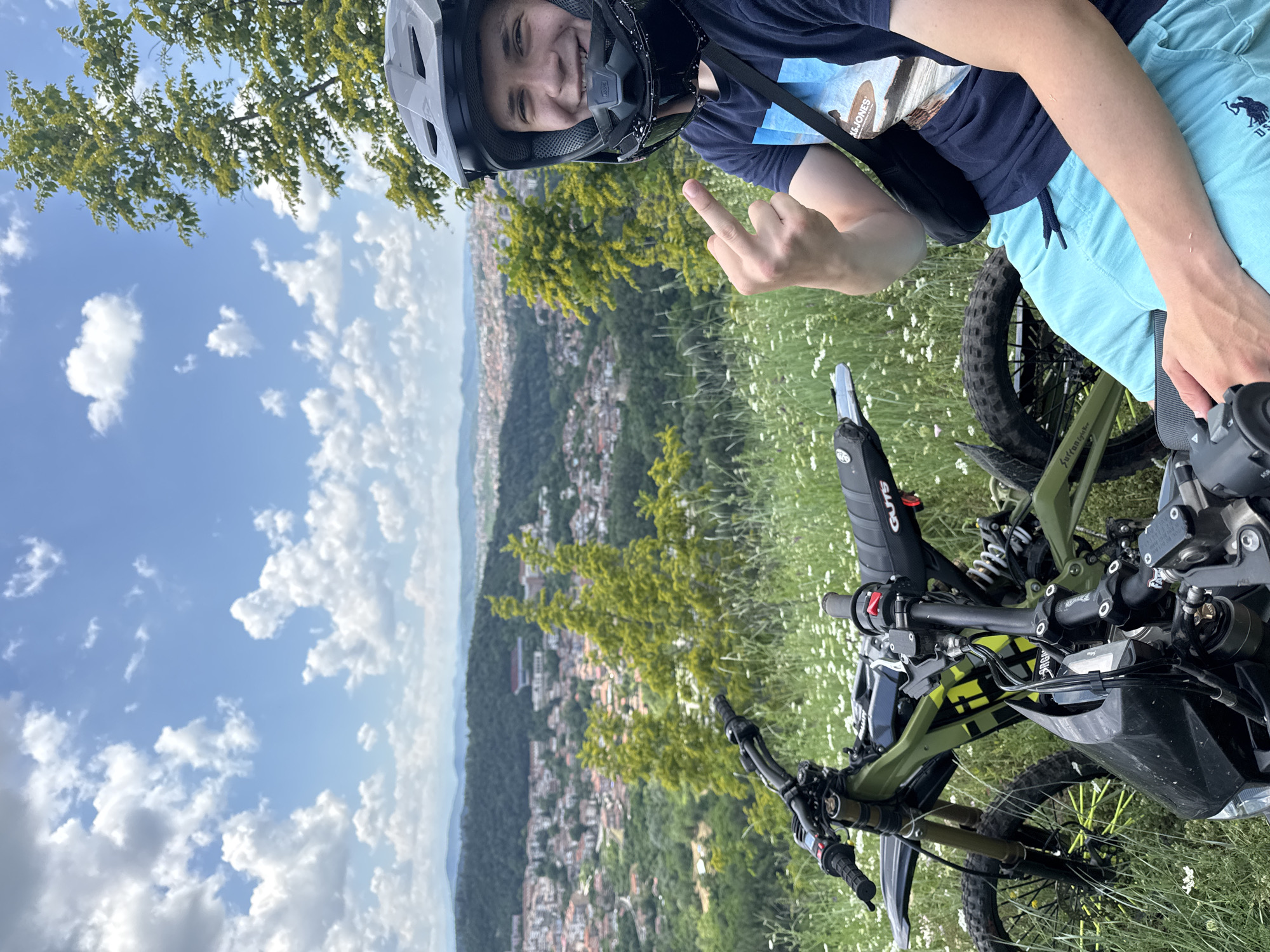 A young Caucasian male wearing a black helmet, dark blue t-shirt, and light blue shorts is sitting on an electric dirt bike. He is smiling and giving the middle finger to the camera. The electric dirt bike is green and black. The background shows a panoramic view of Veliko Tarnovo, Bulgaria, with buildings spread across a hillside. The sky is blue with scattered white clouds. The scene is outdoors, with green grass and small white flowers in the foreground.