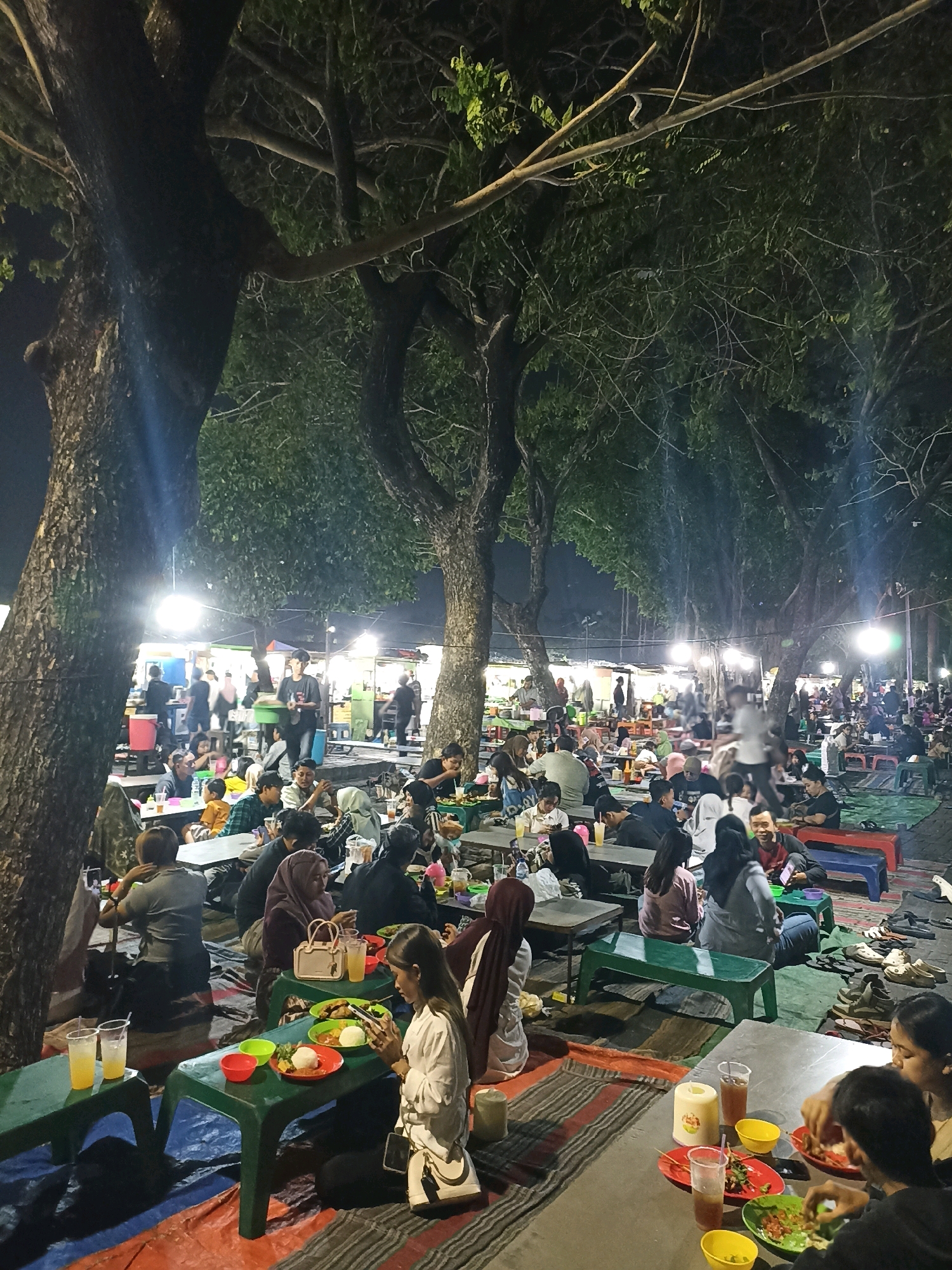 This is a night scene at a bustling outdoor food court in Surabaya, Indonesia, likely near a park or public space given the presence of large trees and open ground covered with mats. Many people are seated at low tables and on the ground, eating and socializing. The atmosphere is illuminated by overhead lights and lights from food stalls in the background. The food court appears to be set up on a paved area, with people utilizing various types of seating, including plastic tables and benches, as well as mats spread on the ground. The overall scene depicts a lively evening gathering, with families and groups of friends enjoying food and company under the shade of trees.