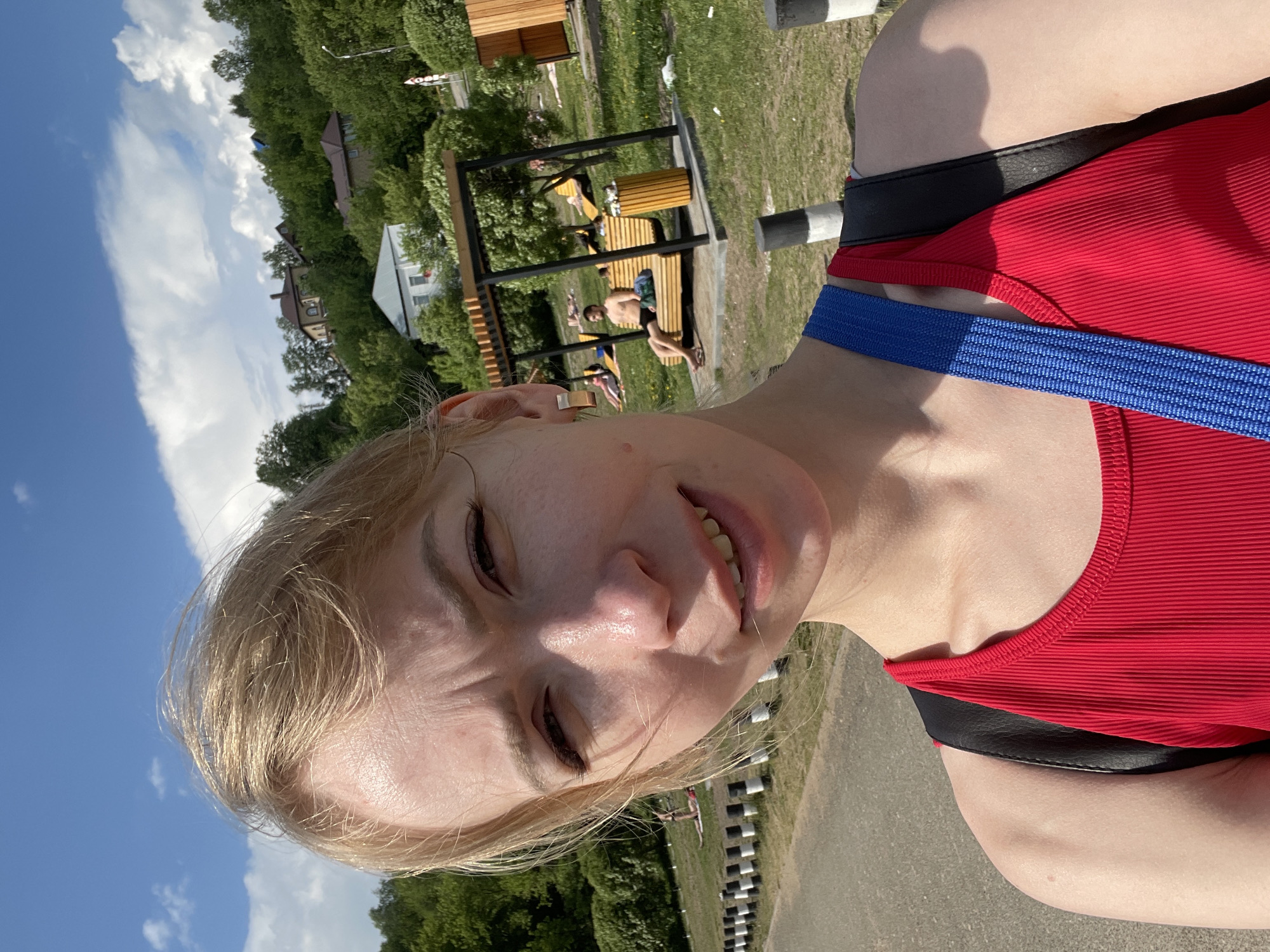A selfie shows a fair-skinned woman with blonde hair. She is wearing a red ribbed tank top with black straps and a blue shoulder strap. The background shows a park-like setting with benches, trees, and other people relaxing. A row of black and white striped barriers lines the foreground. The photo appears to be taken at a slight angle. Based on visual landmarks, the location might be in the city of Perm, Russia, specifically a recreational area such as a park or waterfront promenade.