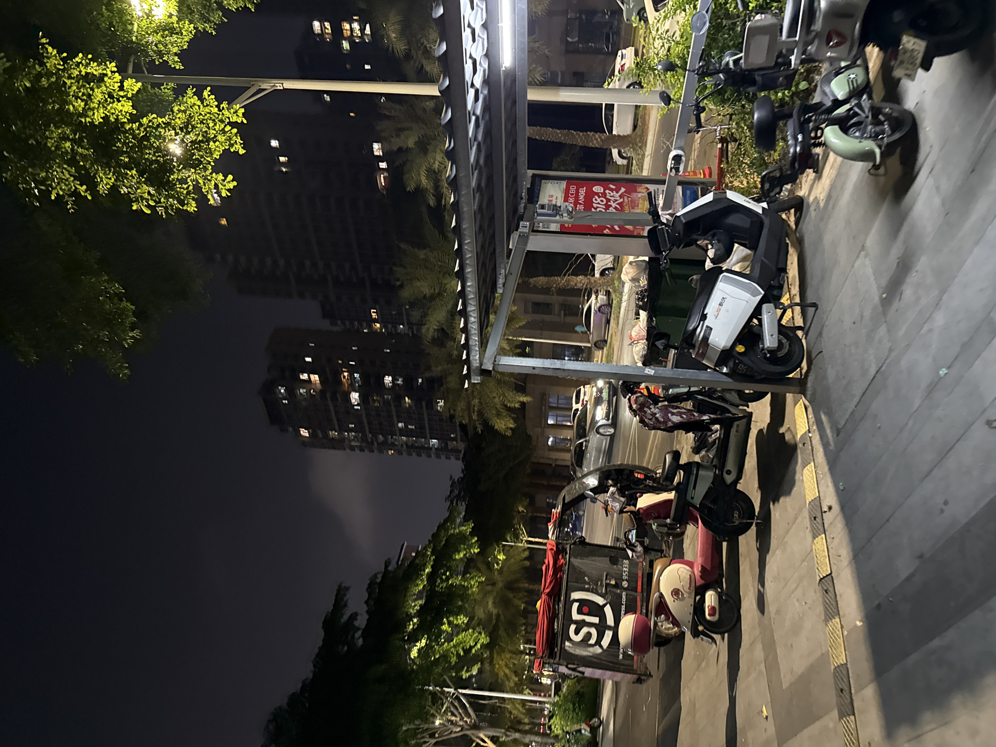 The image depicts a street scene at night in Huizhou, China. In the foreground, several electric scooters are parked. One scooter, a white model, is positioned under a covered structure. To its right, a light green scooter is parked, and partially visible to its right is a light grey scooter. To the left of the white scooter, a pink and cream colored scooter is parked. Further to the left, a delivery scooter with a large black cargo box bearing the "SF" logo is parked. Red fabric is draped over the top of the cargo box. In the background, tall apartment buildings with illuminated windows are visible, interspersed with palm trees. A street lamp with a bright light illuminates the scene from the upper right. Cars are visible on the road behind the scooters. The overall environment suggests an urban setting with a focus on street-level commercial activity and residential areas.