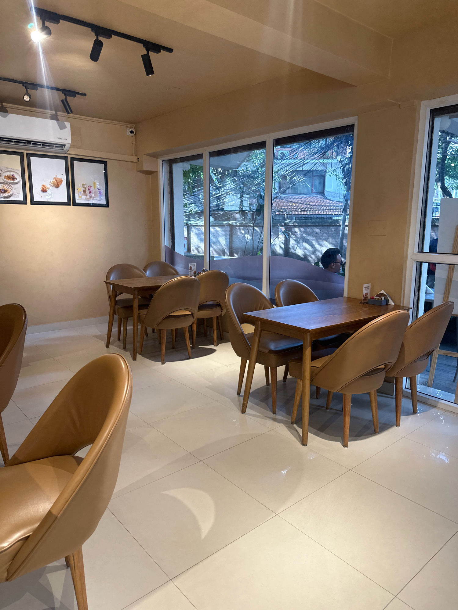 The image depicts the interior of a cafe, likely in Kathmandu, Nepal, featuring a clean and modern aesthetic. Two tables with four chairs each are the central focus, positioned on a light-colored tiled floor. The tables are made of dark wood, and the chairs are upholstered in a warm, tan leather, with wooden legs.
In the background, a large window offers a view of the exterior, revealing trees and buildings, suggesting an urban environment. The walls are painted a muted beige, and track lighting is installed on the ceiling, providing illumination. On one wall, three framed pictures hang, displaying what appear to be food items. An air conditioning unit is also visible on the wall, along with a small security camera. A person is partially visible through the window, their silhouette reflected in the glass. The overall atmosphere is one of a comfortable and inviting dining space.