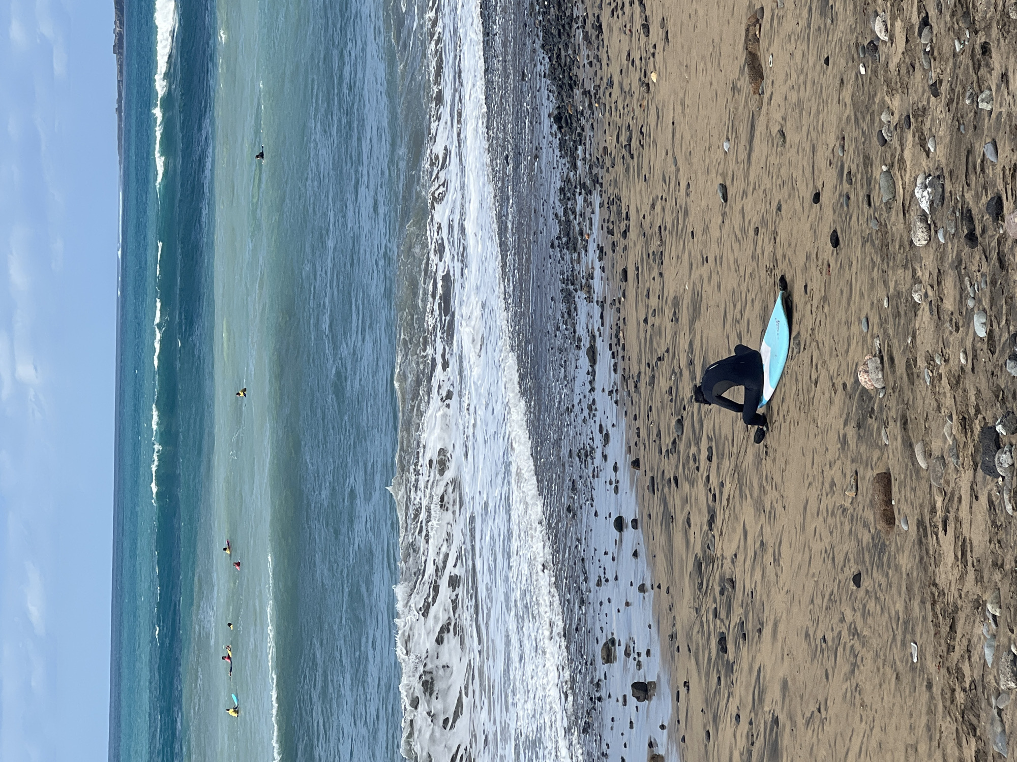 The image depicts a vibrant daytime beach scene in Las Palmas, Spain, centered around surfing. The frame is divided between the ocean on the left and a sandy, pebbly shore on the right.
On the lighter brown sand, which is scattered with various sizes of grey and white pebbles, a person in a dark wetsuit is positioned, leaning over a light blue surfboard. They appear to be in a preparatory stance or resting by the water's edge.
The ocean dominates the left portion of the image, displaying clear blue and greenish hues, indicative of water depth variations. Several breaking waves generate white foam, extending in parallel lines towards the shore. Scattered throughout the water, at least six other individuals in dark wetsuits are visible, many accompanied by surfboards (some with bright yellow or pink highlights). These surfers are either paddling, waiting for waves, or possibly riding them.
The sky above the distant ocean is mostly clear with a few faint clouds, suggesting bright and sunny weather conditions conducive to beach activities. No specific time of day is discernible beyond daylight hours. There is no visible text in the image. The scene conveys an atmosphere of recreation and outdoor leisure.