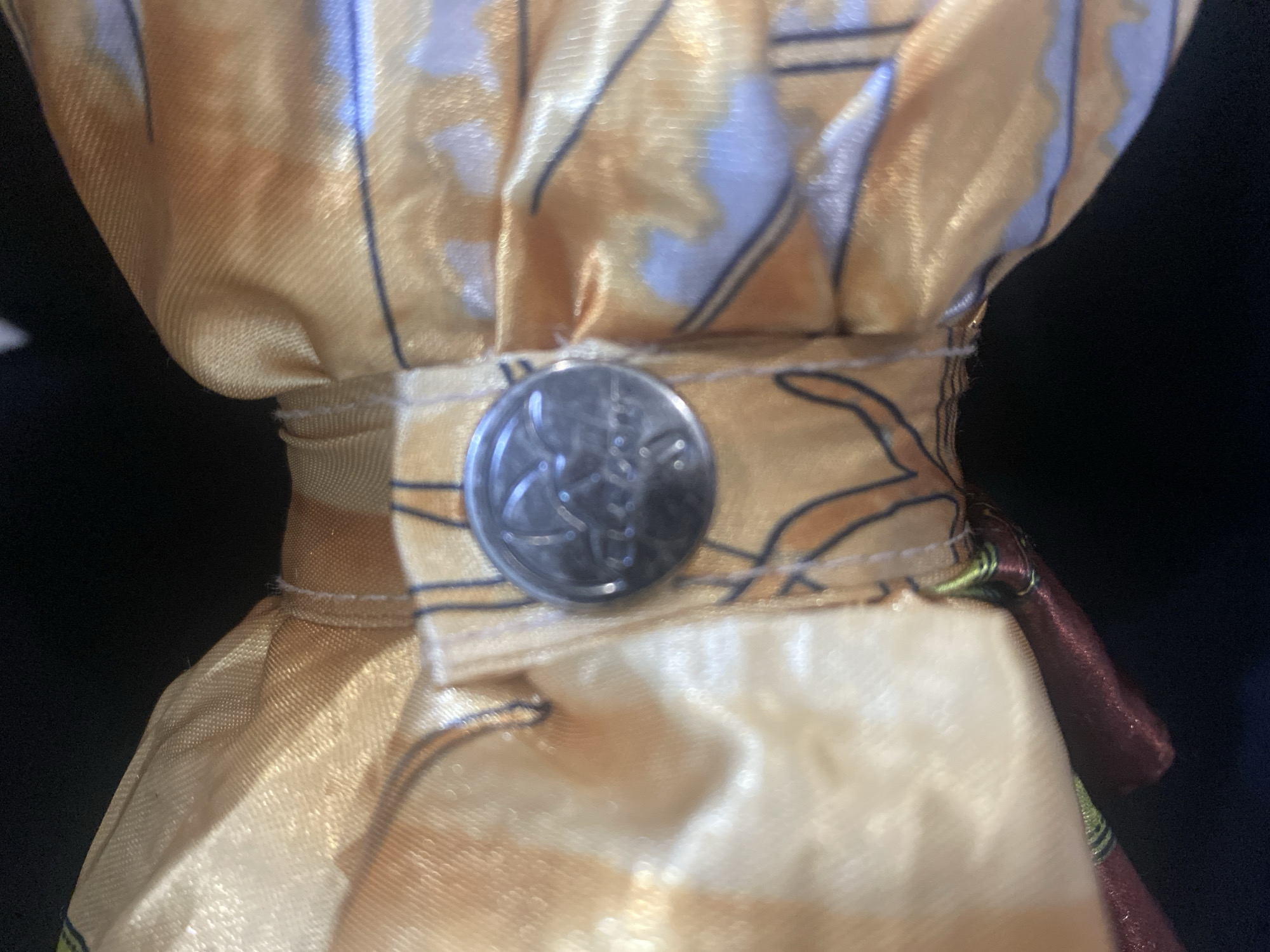 The image shows a close-up view of a golden fabric bow tie, possibly a decorative element or part of an outfit. A circular metal object, resembling a coin or a button with an embossed emblem, is attached to the center of the bow tie. The fabric has a textured, lustrous sheen and is adorned with a pattern of abstract shapes and lines in shades of orange, white, and blue. The bow tie appears to be made of silk or a similar material. The background is dark and out of focus, suggesting the bow tie is either held or laid flat against a dark surface. There is no indication of the location being in Rozhny, Ukraine.