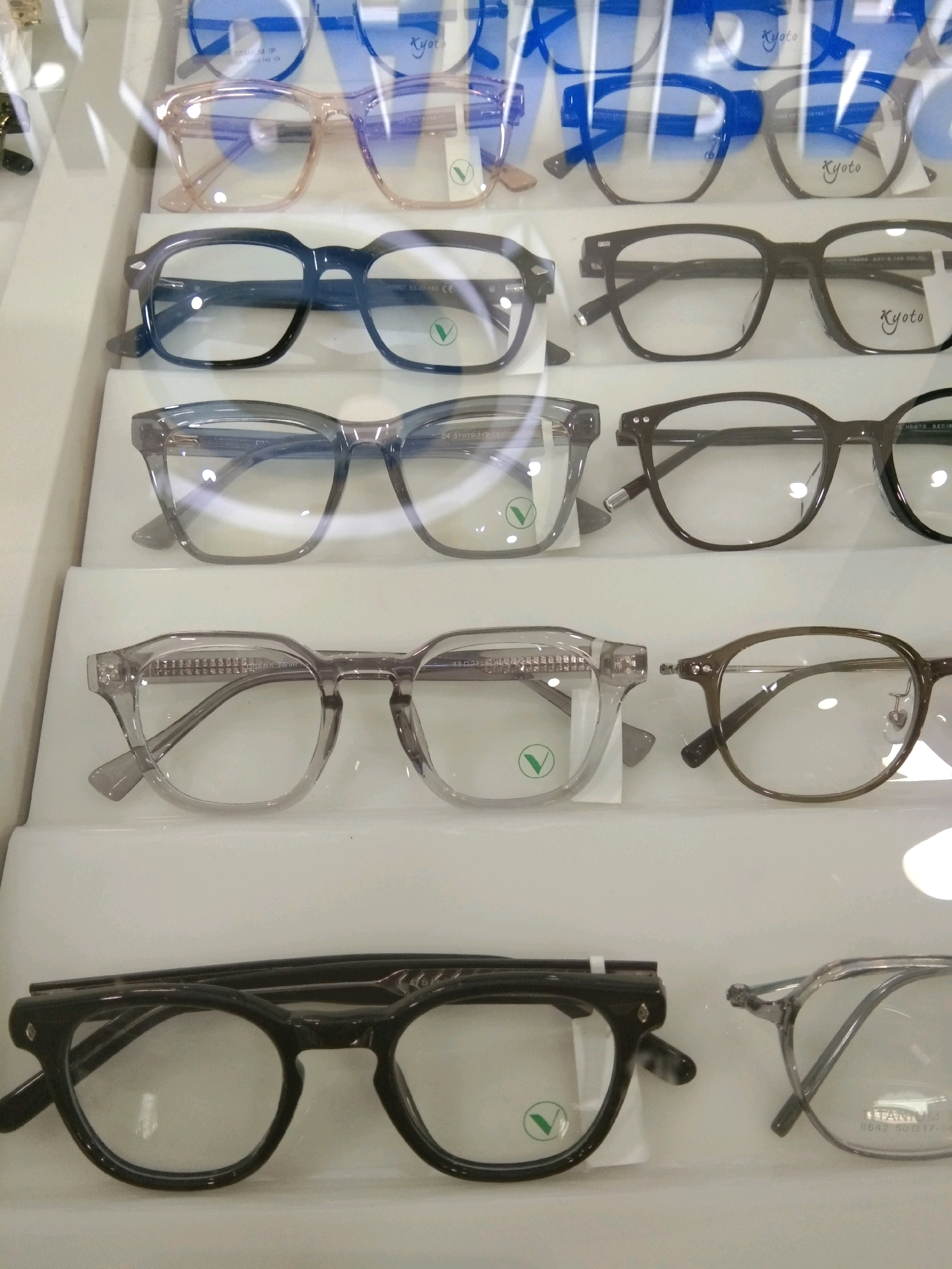 The image displays a selection of eyeglasses arranged on white shelves within a display case. The eyeglasses vary in frame style and color, including black, clear, and translucent grey. Some frames have subtle branding or markings. The lighting suggests an indoor retail environment. The image does not contain any information about the city of Kebumen, Indonesia, or any people.
