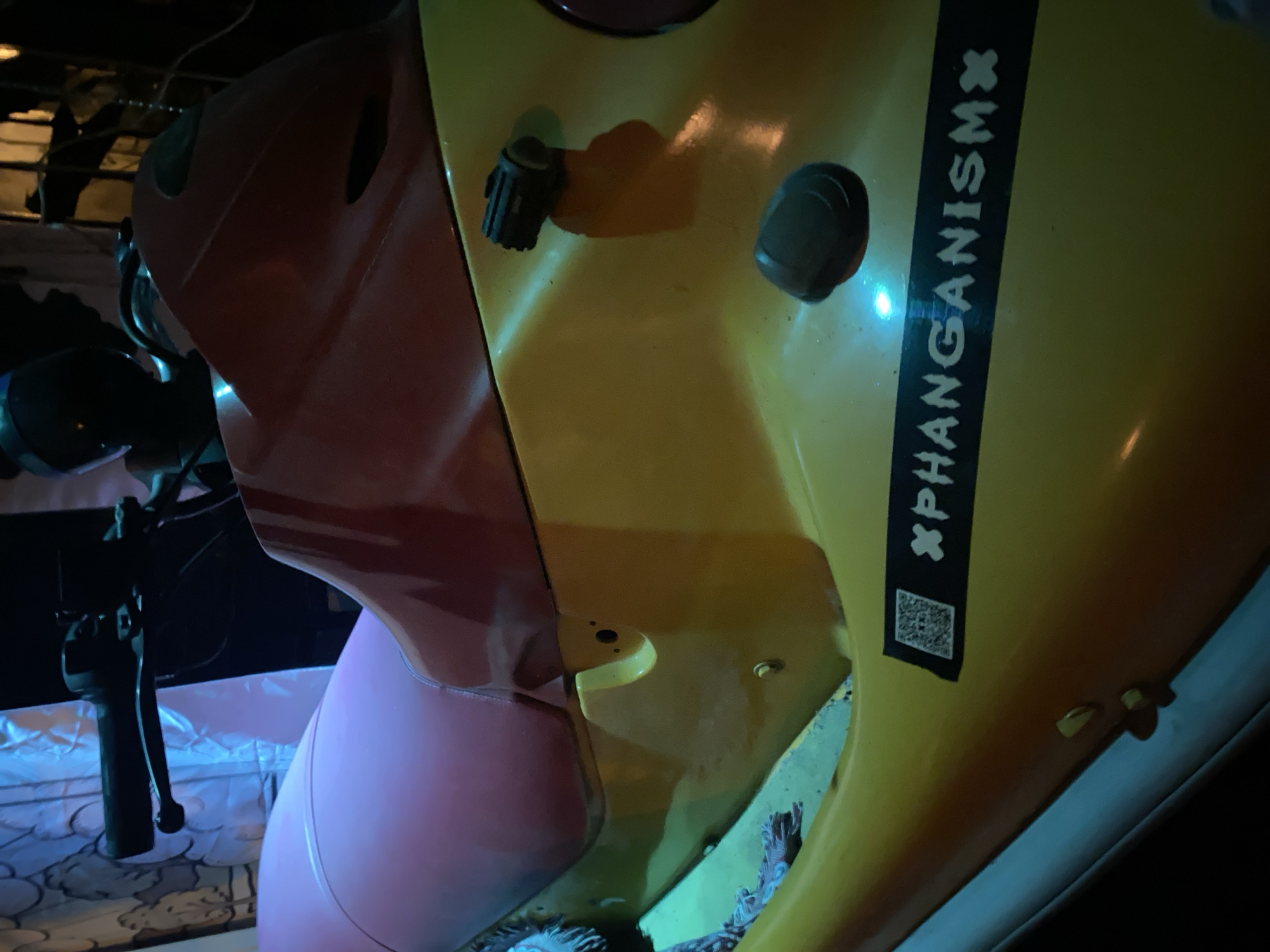 The image depicts a section of a yellow and red vehicle, possibly a customized motorbike or scooter, parked in a dimly lit area. A black sticker with the text "xPHANGANISMx" and a QR code is affixed to the yellow section. The vehicle's handlebars and some mechanical components are partially visible on the left. Based on the text, the location is presumed to be in ตำบลเกาะพะงัน, Thailand.