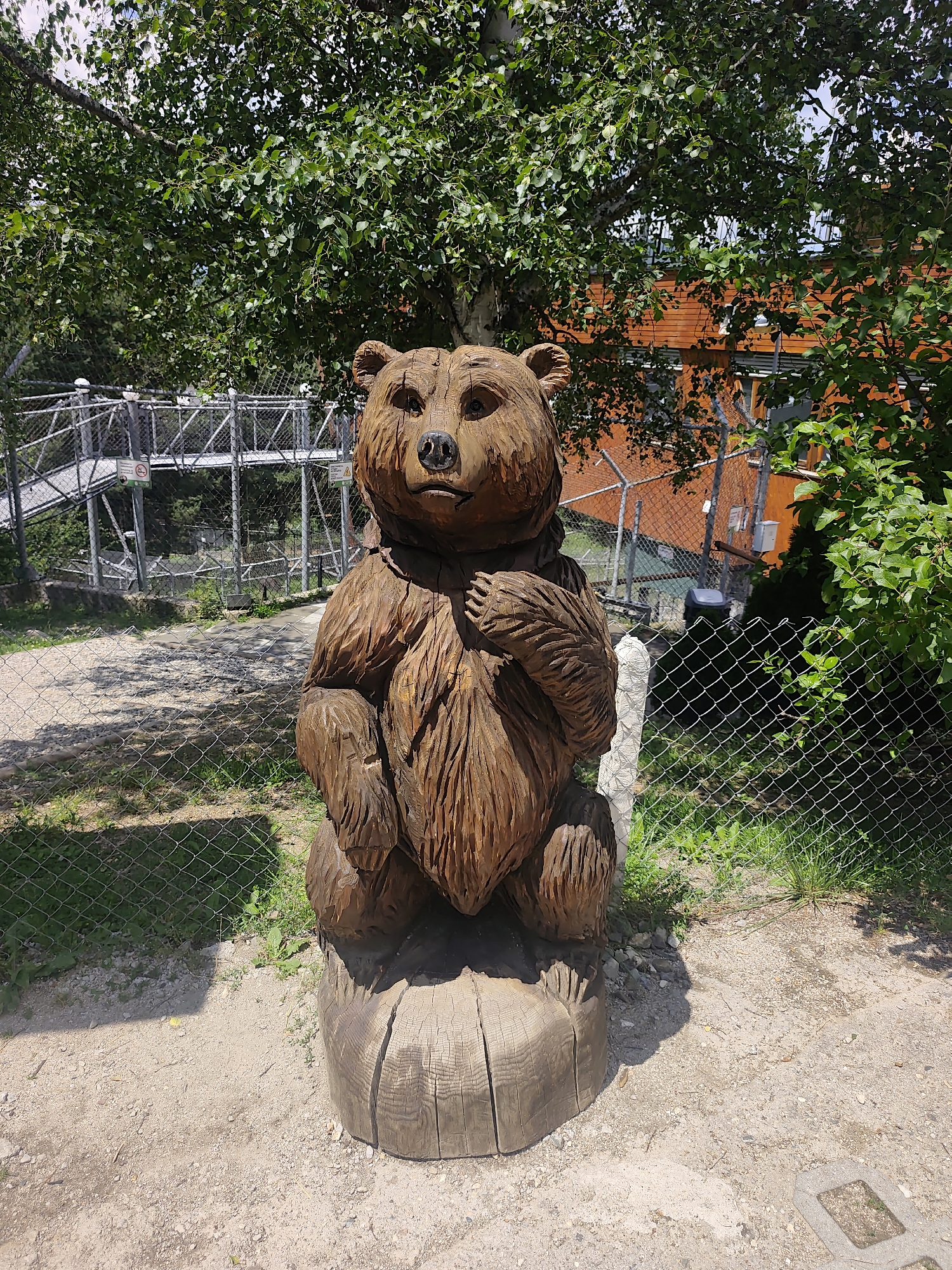 A detailed, full-shot, eye-level, outdoor photograph captures a wooden carving of a bear sitting on a log. The bear carving is brown and textured, with carved fur details and facial features. It is positioned in the foreground, centrally located in the frame. Behind the bear, a chain-link fence runs horizontally. Beyond the fence, a metal walkway or bridge structure is visible, with railings and supports, suggesting an elevated pathway. Further in the background, an orange building with windows can be seen, partially obscured by lush green trees and foliage. The ground in front of the bear is a mixture of sandy soil and small pebbles. The overall environment suggests an outdoor recreational area, possibly a park or a zoo exhibit, in the city of Yakoruda, Bulgaria. The lighting indicates a bright, sunny day with shadows cast on the ground.