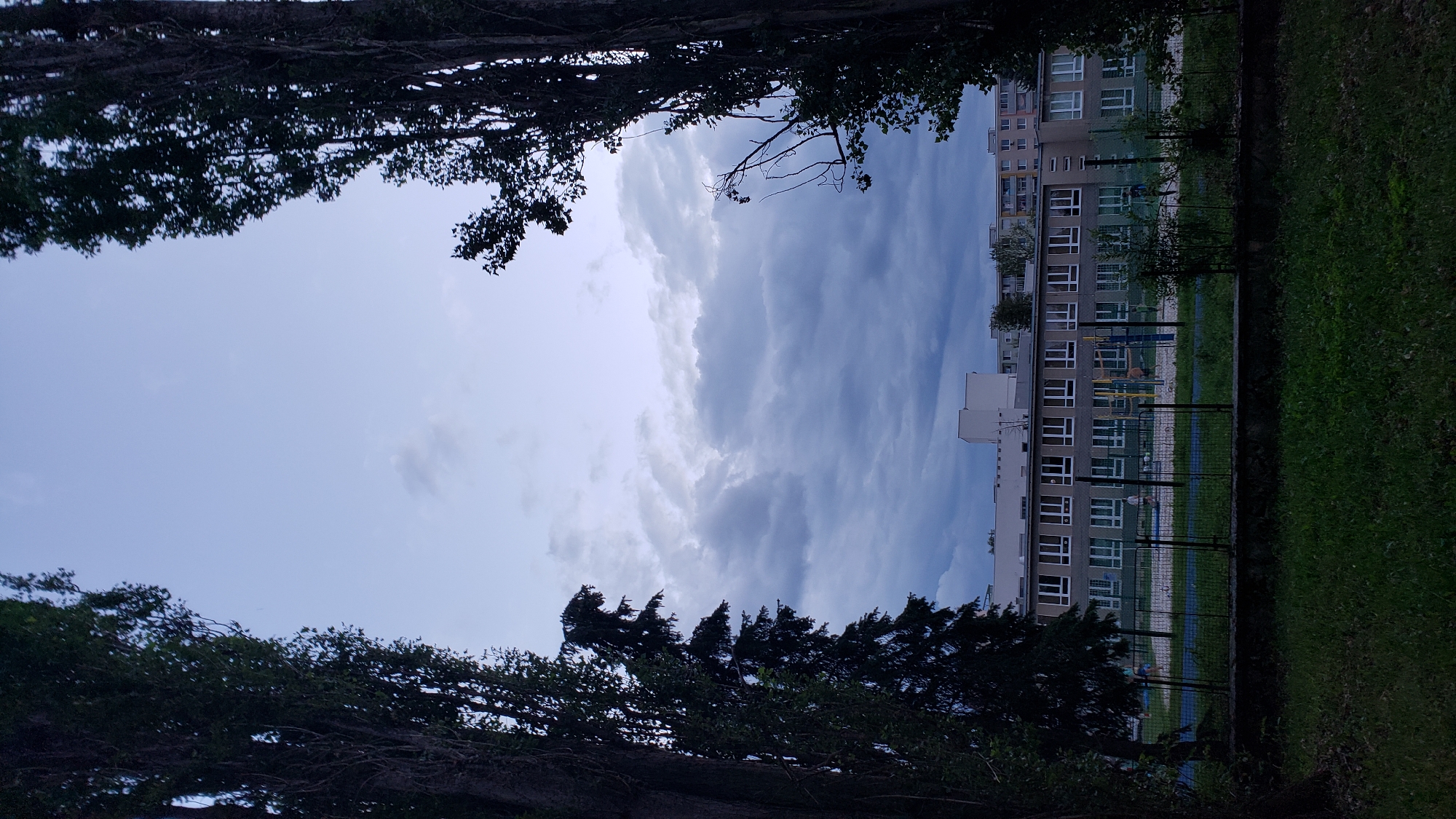 The image shows a view of a school building from across a grassy area and a fence. Two tall, dark trees frame the sides of the image, creating a natural tunnel effect. The sky above is overcast with dark, heavy clouds, suggesting an impending storm or the end of the day. The school building appears to be multi-story, with a facade of windows. In front of the building, there's a fenced-in playground area with some playground equipment. A few people are visible on the playground, appearing to be children and possibly adults supervising. The overall scene is somewhat somber due to the dark sky and the overcast conditions. The location is identified as being in Prešov, Slovakia.