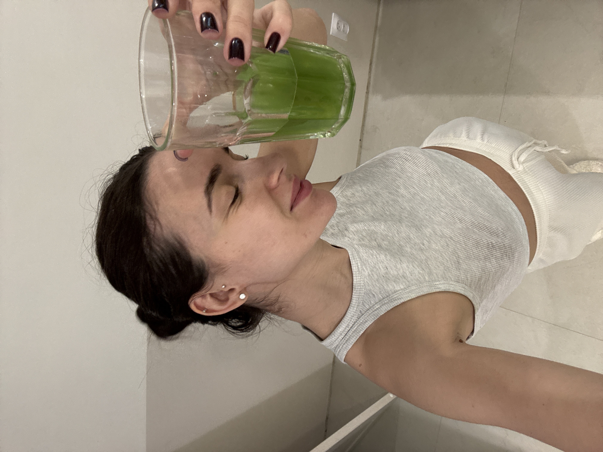 A woman is holding a glass of green liquid above her head. She is wearing a light grey tank top and white pants. She has dark hair pulled back. Her nails are painted dark purple. The background appears to be a kitchen or bathroom with white cabinets and a tiled floor. The specific location is not identifiable from the image.