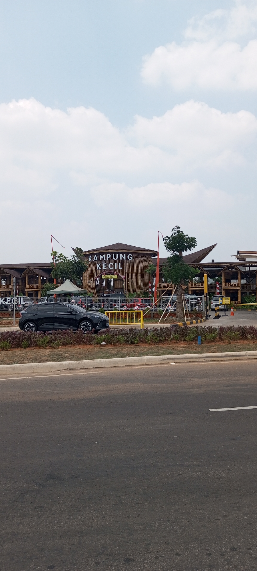 The image depicts the exterior of a building complex with a prominent sign that reads "KAMPUNG KECIL". The architecture features a unique design, with a large circular structure made of bamboo prominently displayed. The building appears to be a restaurant or a leisure facility, as suggested by the presence of tables and chairs visible through the open structures. In front of the building, a black SUV is parked on a paved area, with a yellow barrier separating it from the road. The scene is set under a cloudy sky. The location is identified as Rawa Banteng, Indonesia.