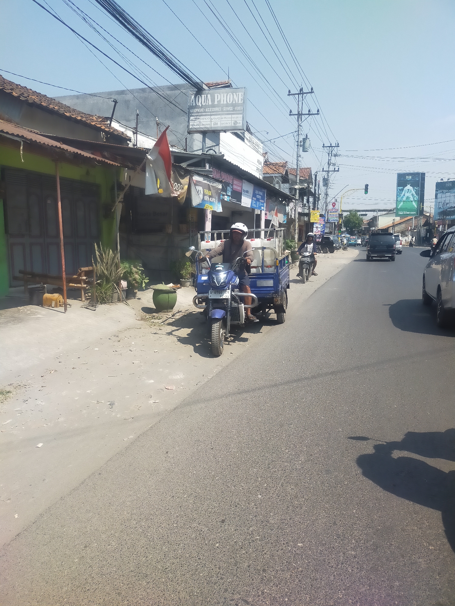 The image shows a street scene in Talang, Indonesia. A blue three-wheeled motorcycle with a cargo bed is parked on the left side of the road, near a sidewalk. A person wearing a helmet is seated on the motorcycle, appearing to be the driver. The cargo bed contains several white plastic containers. The motorcycle has a license plate that reads "G 6966 CY".

To the left of the motorcycle, there are shops with awnings and closed doors. A flag with red and white stripes is visible on one of the buildings. A green planter pot and some plants are also on the sidewalk. In the background, there are power lines and utility poles. Further down the street, other vehicles, including cars and motorcycles, are visible. There are also several billboards and signs, one of which advertises "AQUA PHONE". The road is asphalt and appears to be in good condition. The sky is clear and blue. The scene is illuminated by sunlight, casting shadows on the road. The overall impression is a typical street scene in a commercial area of a town.