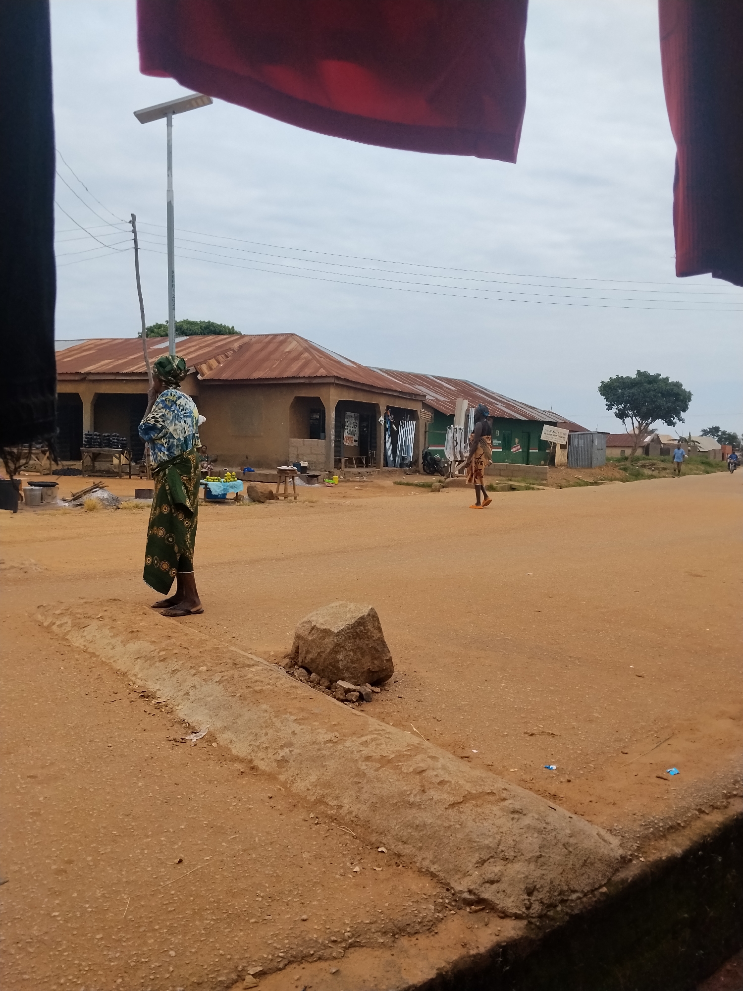 An outdoor scene in Mangu Town, Nigeria, displays a dirt road with buildings along the side. In the foreground, a woman stands facing away from the camera, wearing a patterned headscarf and a long, patterned skirt. The road is lined with shops that have corrugated metal roofs. A solar-powered street lamp is visible in the background. Further down the road, another woman walks away from the camera. The sky is overcast.