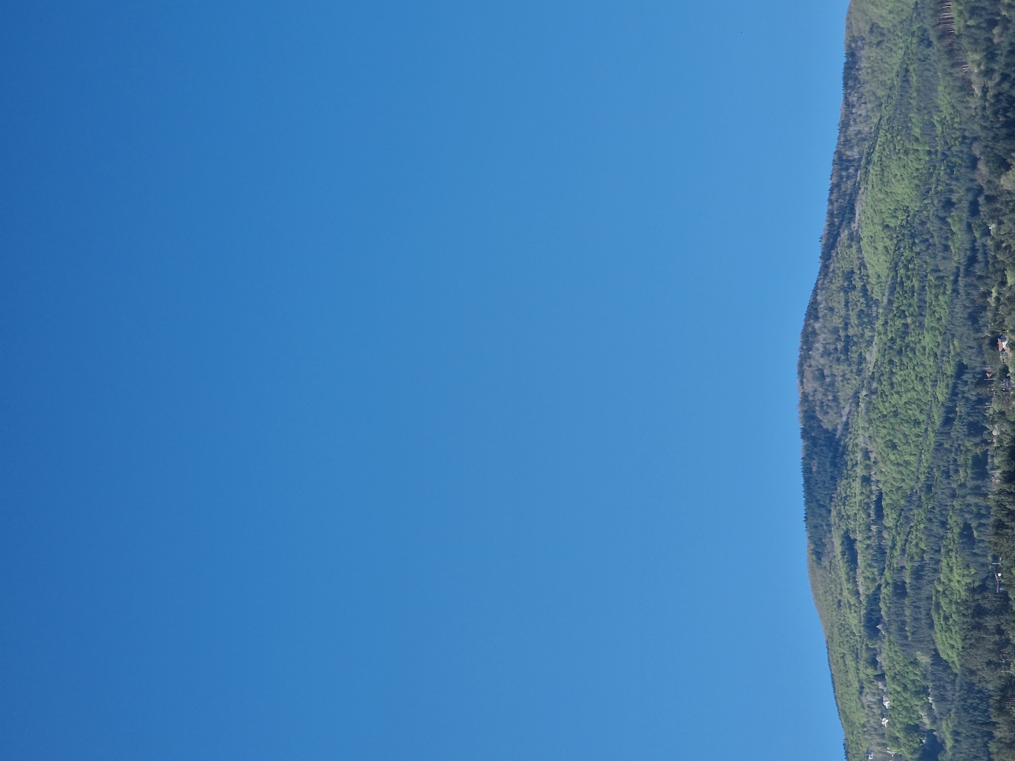 The image shows a clear, blue sky taking up most of the frame. A mountain covered in lush, green vegetation occupies the right edge of the image. The location is Unknown, Unknown.