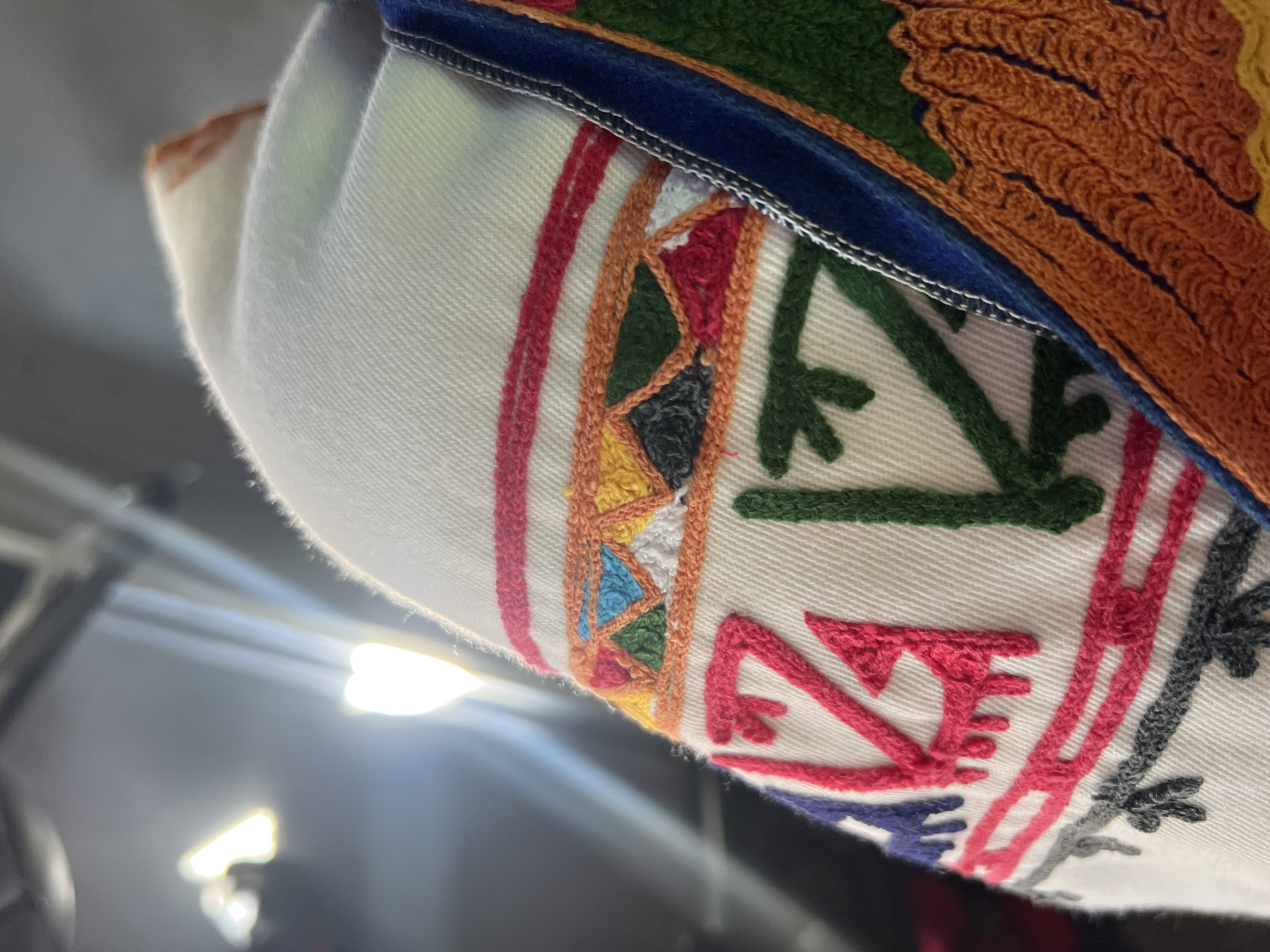 The image displays a detailed, close-up view of a white textile adorned with elaborate, multi-colored embroidery, situated in Slivovo, Bulgaria. The base fabric is an off-white material with a discernible weave pattern. The embroidery is executed with vibrant threads in numerous colors including bright pink/magenta, dark green, blue, orange, yellow, red, and dark grey.
Two main horizontal bands of embroidery are prominent. The upper band comprises a series of geometric shapes such as triangles and squares, filled with dense, textured stitching in varying hues including yellow, orange, green, blue, white, black, and red. Below this, distinct stylized angular motifs are stitched directly onto the white fabric; a prominent pink/magenta design resembling a stylized letter or symbol is located on the left, positioned next to a green angular pattern. Further down, narrower horizontal bands feature small, repeating decorative elements in pink/magenta and dark grey.
A vertical dark blue trim, possibly velvet, runs along the right edge of the embroidered white textile, separated by a fine white stitching line. Adjacent to this blue trim, an area of rich orange embroidery displays a wavy or scalloped pattern. The background is out of focus and appears dark, containing several bright, blurred light sources in the upper left quadrant. One light source exhibits a distinct heart shape, contributing to the bokeh effect. The lighting suggests an indoor setting. No individuals or specific actions are discernible within the frame.