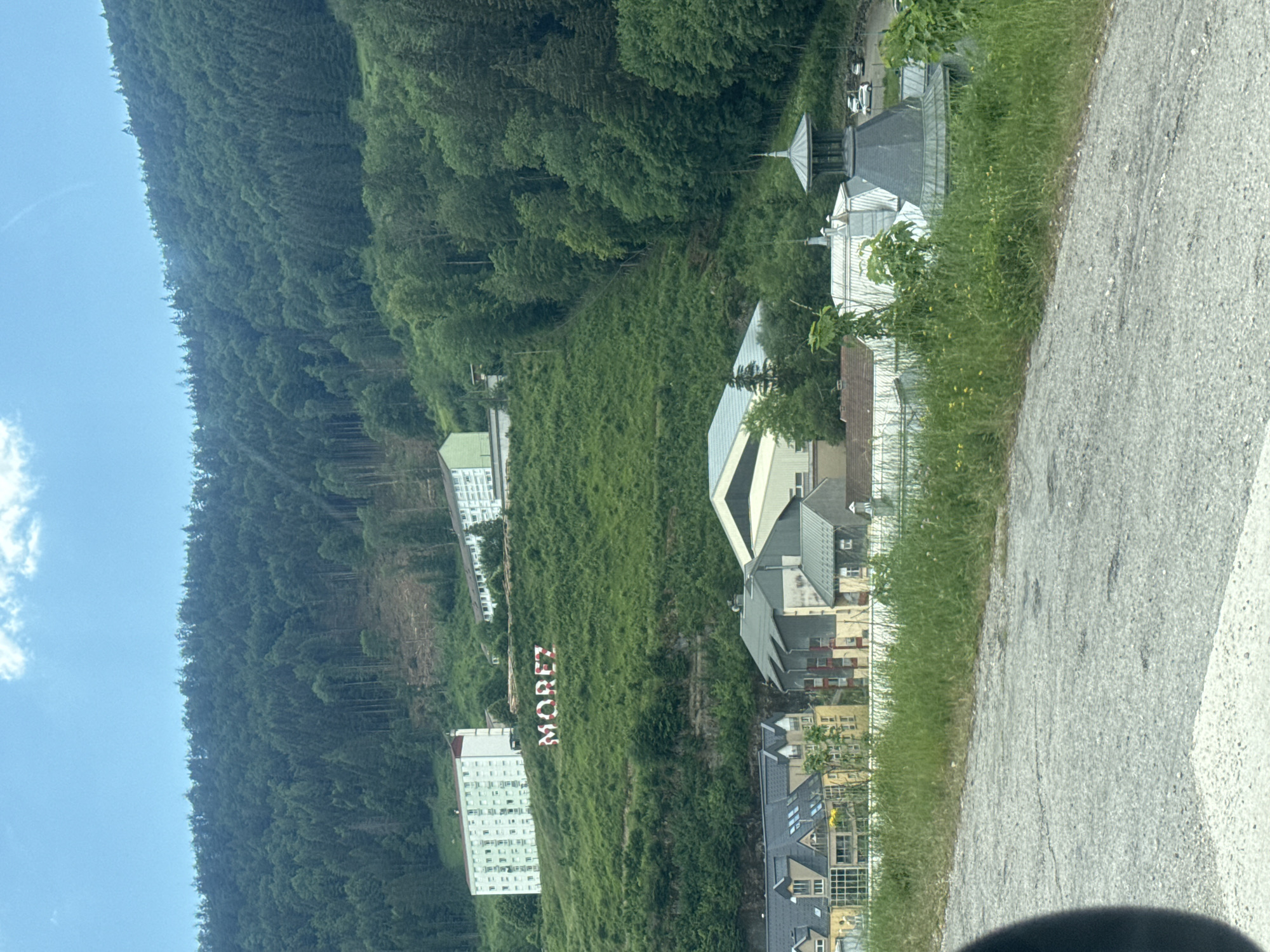 The image shows a hillside with the word "MOREZ" spelled out in large, red and white letters on the grassy slope. Two multi-story white buildings are situated higher up on the hill, one with a red roof and balconies, and another with a white roof. Below the large letters, several buildings are visible, including structures with multi-pitched roofs and a building with a distinctive grey, domed cupola. The overall setting is mountainous and forested, with a cloudy blue sky overhead. The scene appears to be in the town of Morez, which is now part of the city of Hauts de Bienne in France.
