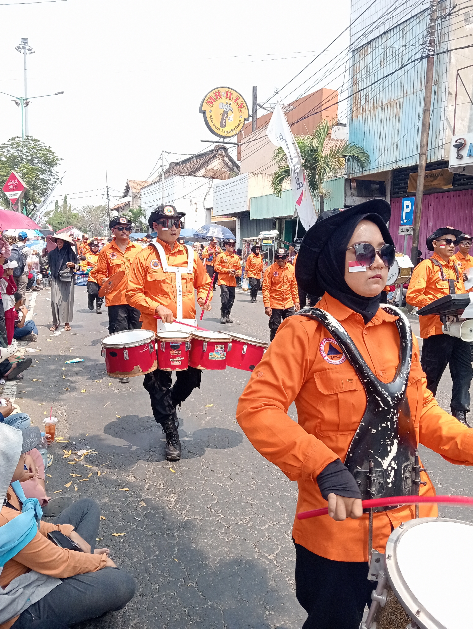 A group of people in orange uniforms and black hats with sunglasses are marching in a parade on a paved street in Demak, Indonesia. The person in the foreground is a woman wearing a black hijab and sunglasses, with red and white face paint on her cheeks. She is holding red drumsticks and appears to be playing a drum. Other individuals in the parade are also carrying drums and wearing similar uniforms. Onlookers are seated on the side of the street, some holding drinks. Buildings line the street, with shops and signs visible. A "MR. D.I.Y." sign is prominent on one building. The sky is bright and clear, suggesting it is daytime. The scene depicts a festive or ceremonial event within the city.
