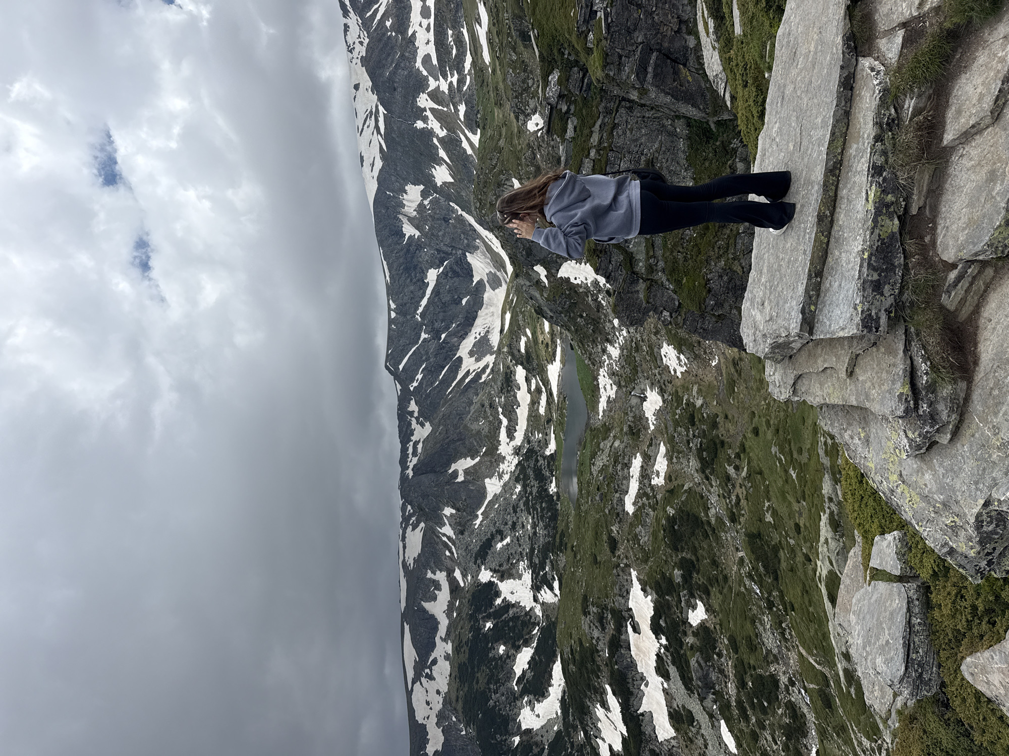 A woman stands on a rocky outcrop overlooking a mountainous landscape in Panichishte, Bulgaria. The woman is facing away from the camera, with her hands near her face as if shielding her eyes from the sun or shielding herself from the wind. She is wearing a grey hooded sweatshirt and black pants. The landscape features rugged, snow-dusted mountains under a cloudy sky. A small lake is visible in the valley below, surrounded by green vegetation and rocky terrain. The rocks in the foreground are large and irregular, with patches of moss and grass. The overall scene depicts a remote, natural environment.