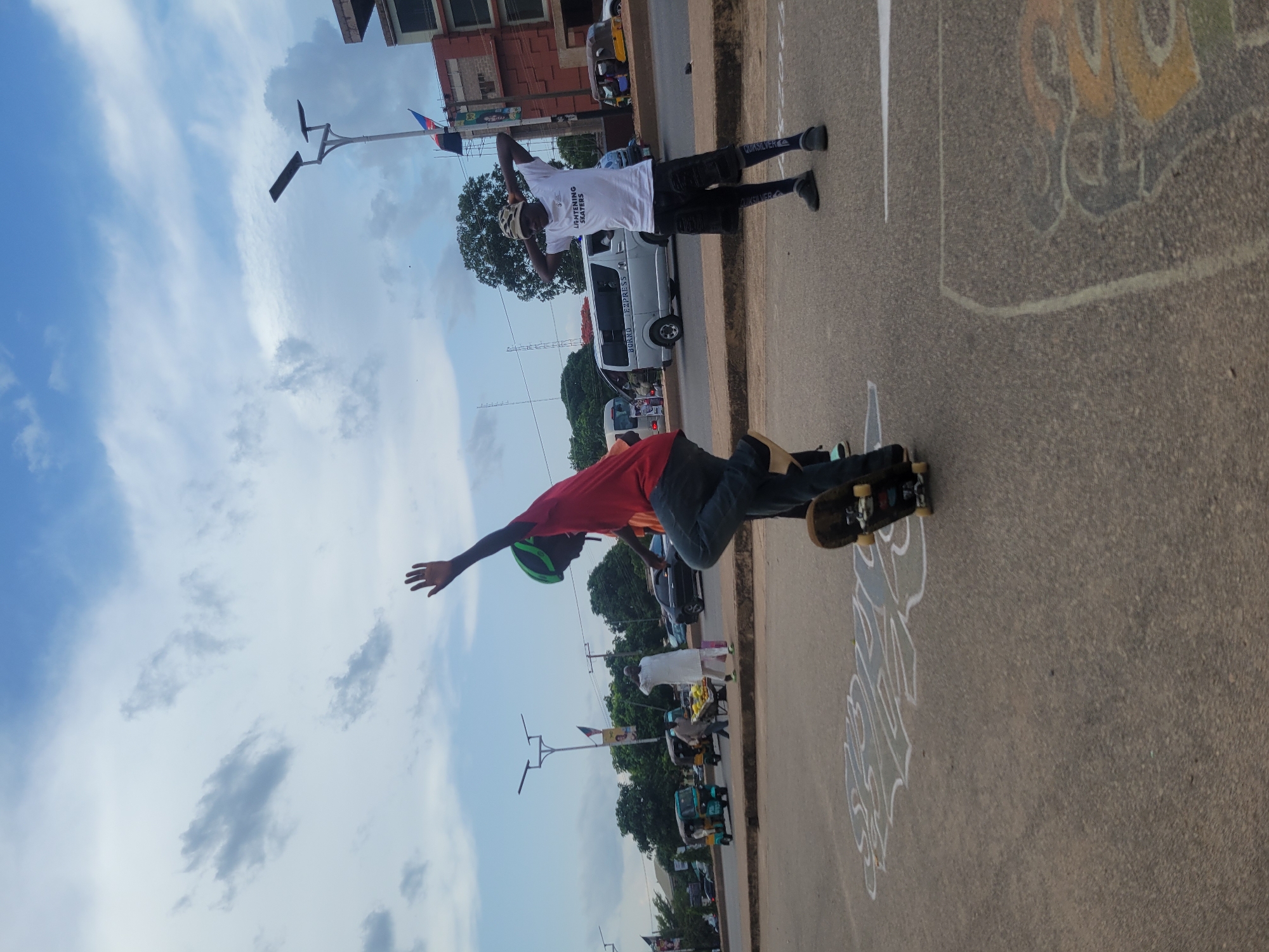 The image depicts an outdoor scene in Jos, Nigeria. A young person in a red shirt, dark pants, and a green helmet is riding a skateboard on a paved road. Another individual stands nearby wearing a white shirt and dark shorts. The road features painted artwork. In the background, there is a white van, other vehicles, trees, and streetlights under a partly cloudy sky. Buildings are visible in the distance.