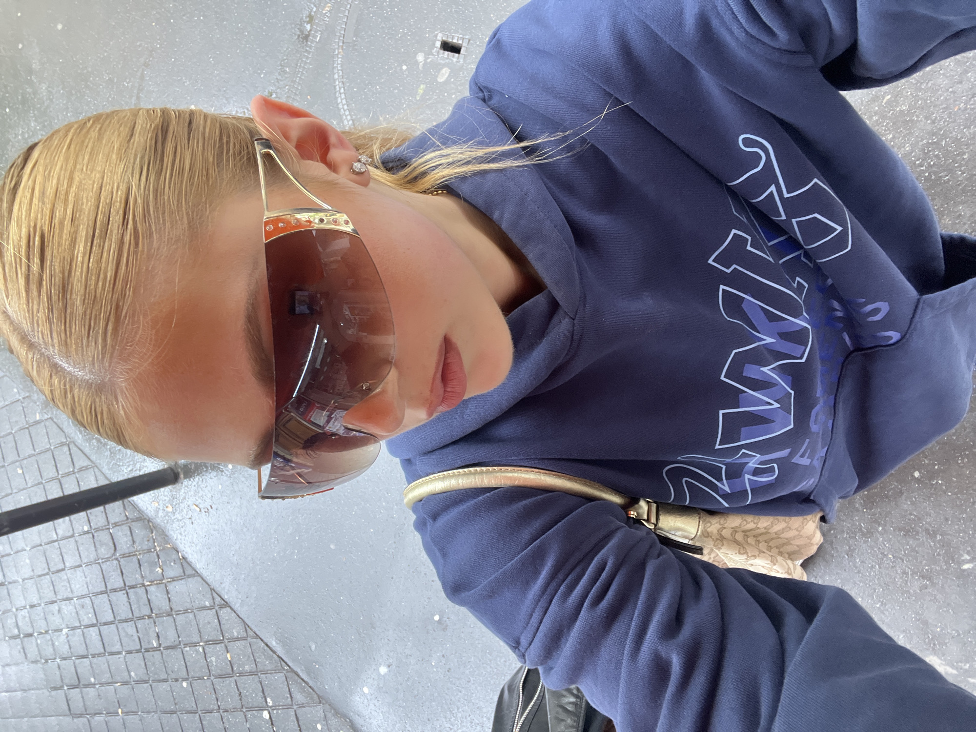 A woman with blonde hair, wearing oversized brown sunglasses and a navy blue hoodie with white lettering, is standing on a gray tiled surface in what appears to be an urban environment. A gold purse strap is visible across her torso. The overall setting suggests an outdoor location in a modern city. The precise location within Paris is not discernible from the image.
