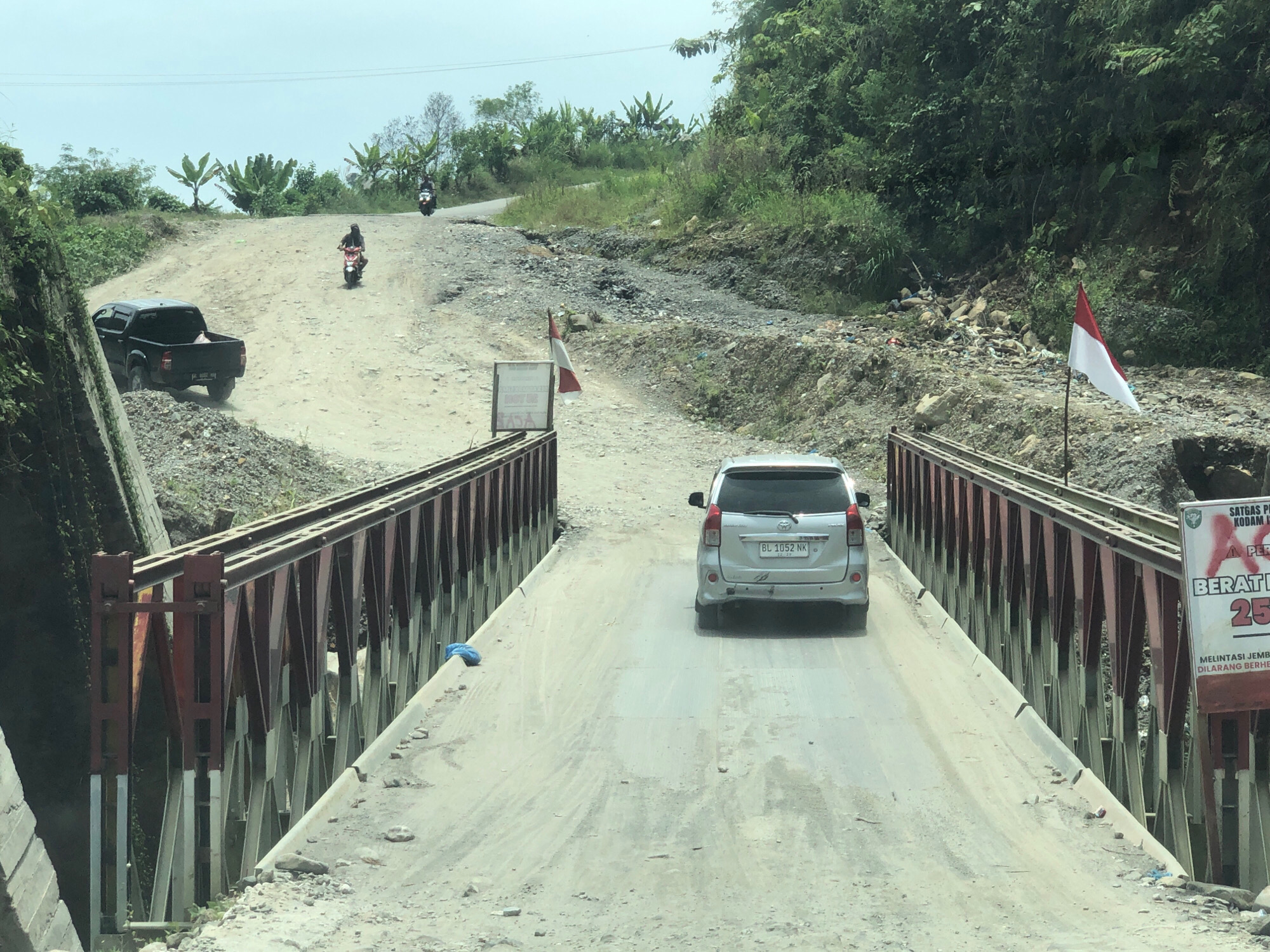 In the city of Bintangpermata, Indonesia, a red and white Indonesian flag flutters on a pole beside a sign that reads "SATGAS PKB KODAM..." and "BERAT MAX 25 TON". A silver minivan with license plate "BL 1052 NK" drives across a bridge. The bridge itself is made of metal with red and silver supports and has a dirt road surface. The surroundings are hilly with dense green vegetation and some rocky, eroded terrain. A black pickup truck is parked on a dirt road in the distance, and two individuals on motorcycles are also visible on the road ahead.