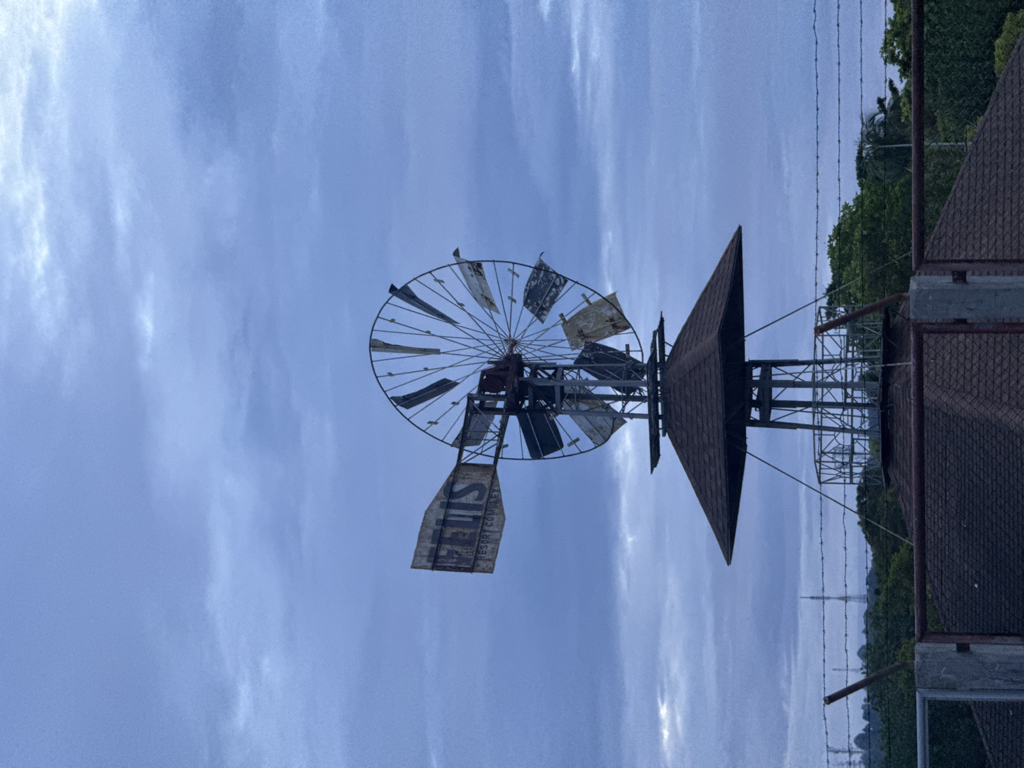 The image shows a rusted and deteriorated windmill structure located in Davao City, Philippines. The windmill is positioned against a cloudy blue sky. The blades of the windmill are incomplete and some are missing. An advertisement sign is attached to the structure. The windmill is near buildings with brown shingle roofs and trees. A barbed wire fence is visible between the windmill and trees.