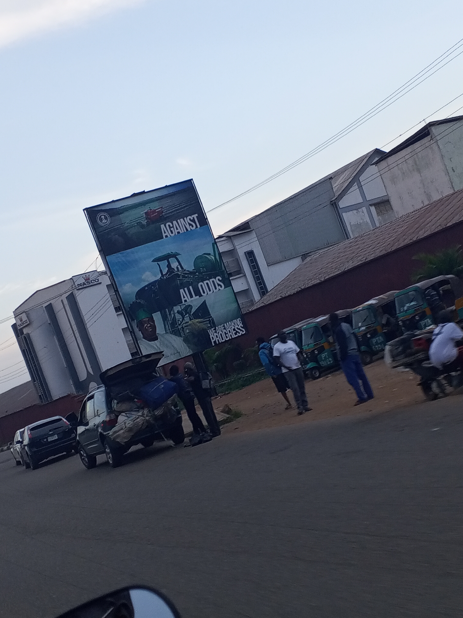 In Jos, Nigeria, a billboard with the words "AGAINST ALL ODDS" is displayed. A man in a green hat is featured on the billboard, along with images of tractors. Several people stand near the billboard. In the foreground, a dark-colored SUV with its trunk open and luggage piled high is parked. Behind it, a black car is also visible on the road. Several three-wheeled vehicles, possibly auto-rickshaws or minibuses, are lined up on the side of the road, and a motorcycle is parked nearby. The scene appears to be a busy roadside area with commercial buildings in the background.