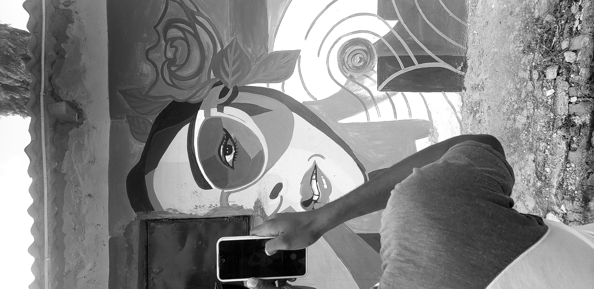 In the city of Narayi, Nigeria, a person's hand holds a white smartphone, capturing an image of a vibrant mural on a weathered wall. The mural depicts a woman with large, fashionable sunglasses and a rose adorning her hair, her face rendered in bold, graphic lines and contrasting shades of gray. The wall itself shows signs of age and neglect, with peeling paint and a rough texture. In the foreground, the person's arm and shoulder are visible, clad in a dark shirt. The ground below is uneven, covered in rubble and debris, suggesting an urban setting that is perhaps undergoing development or repair. The overall scene is a snapshot of everyday life, where modern technology intersects with street art and the realities of the urban environment.