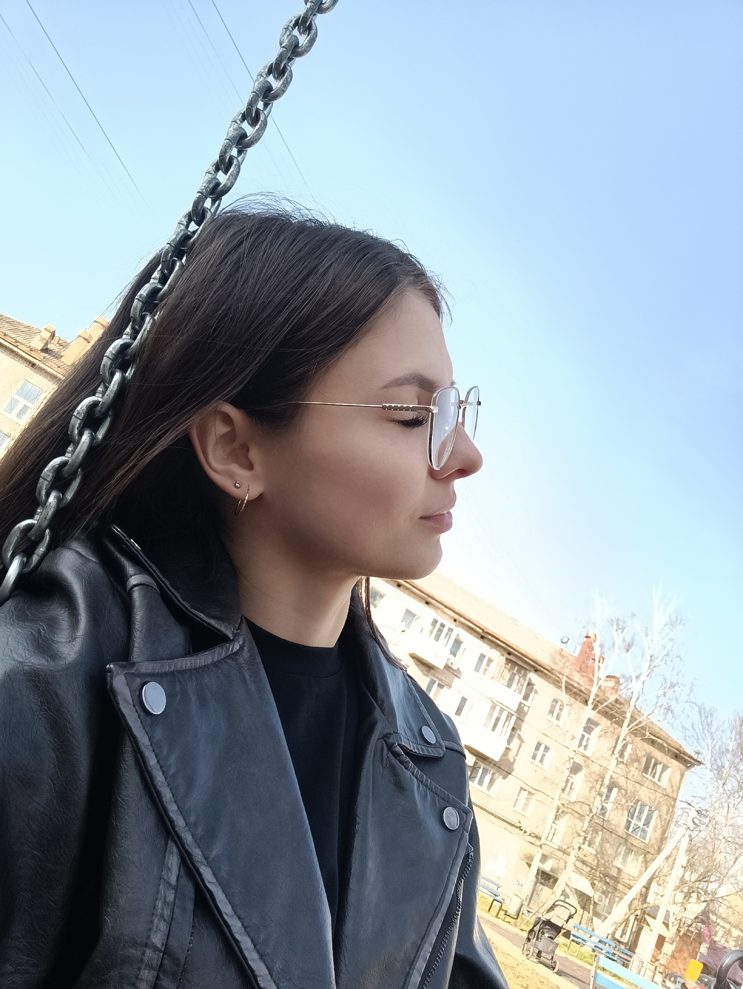 A medium, outdoor shot depicts a young, white female wearing a black leather jacket, a black t-shirt, and gold-rimmed glasses. She has long, dark brown hair. She is wearing gold hoop earrings and has at least one piercing in her ear. The female is in profile, looking right. A thick, steel chain is visible on the left. Behind her, there is a light blue sky with thin power lines. In the background is an apartment building with light colored walls and numerous windows. A tree with bare branches is to the right of the building. Further down and to the right is a playground with blue railings and a black stroller. The setting seems to be a city park in an urban environment. The city and region are unspecified.