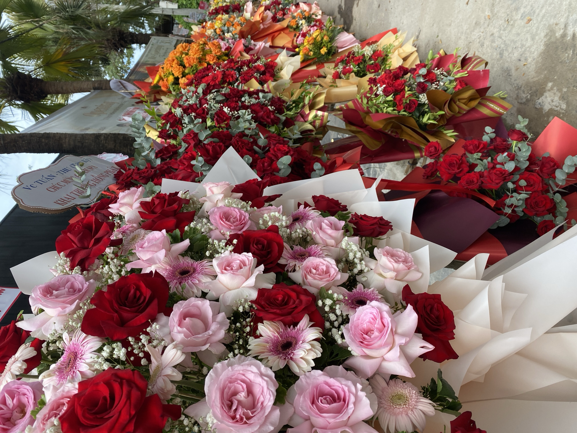 The image shows a row of large floral arrangements, likely for a grand opening or celebration, displayed outdoors. The arrangements feature a variety of flowers including roses and gerbera daisies in shades of red, pink, and orange, interspersed with greenery and small white flowers. Some of the arrangements are adorned with decorative ribbons and wrapping. In the background, a large sign with Vietnamese text is visible, along with palm trees and a wall. The scene is likely located in a city in Vietnam, possibly on a street or sidewalk, given the presence of pavement and the outdoor setting. The style of the floral arrangements and the signage suggest a festive or commercial event.